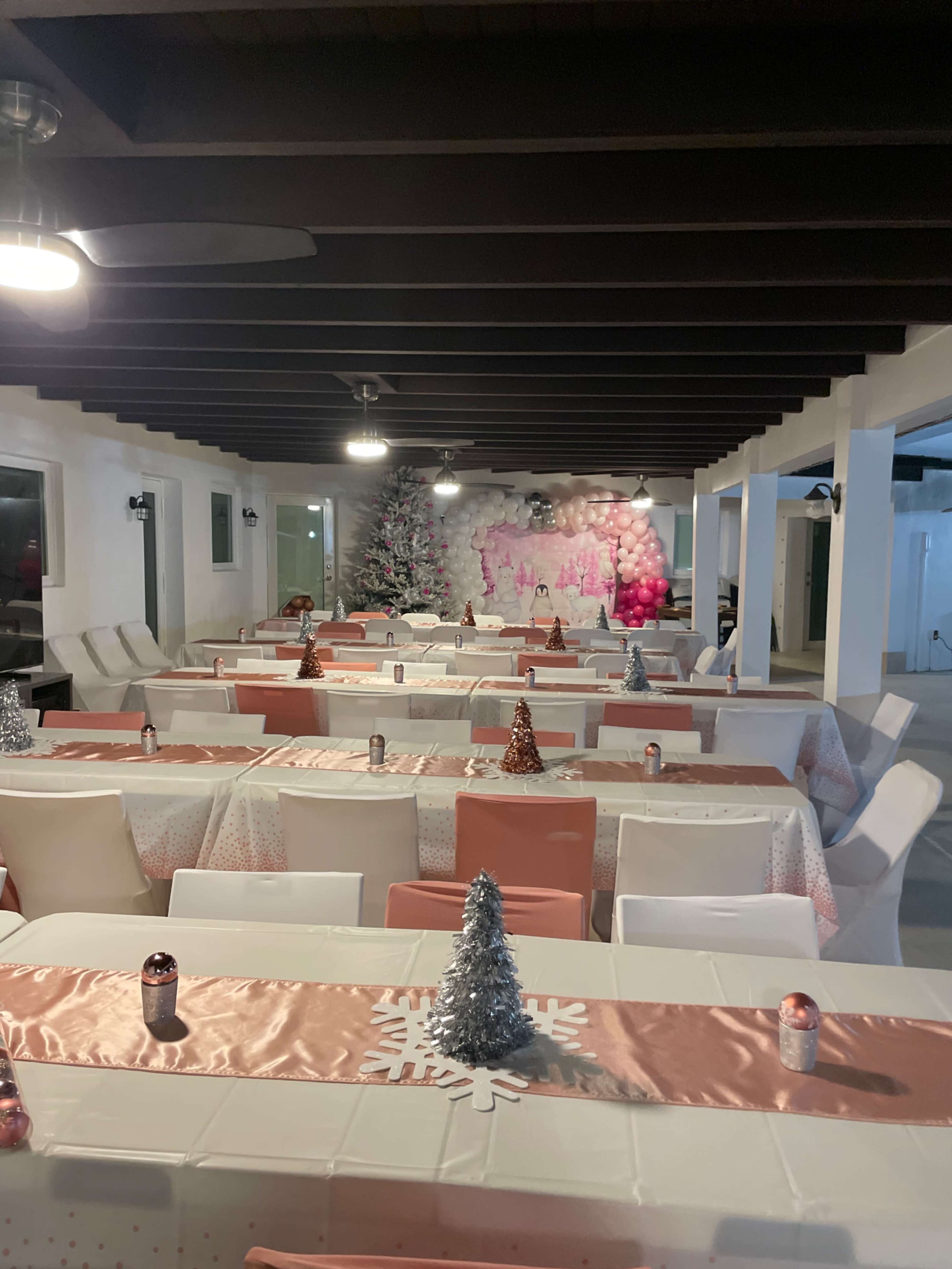 A spacious event hall is set up with tables dressed in white and pink tablecloths decorated with small Christmas trees and candles, under a ceiling with exposed beams and overhead fans.