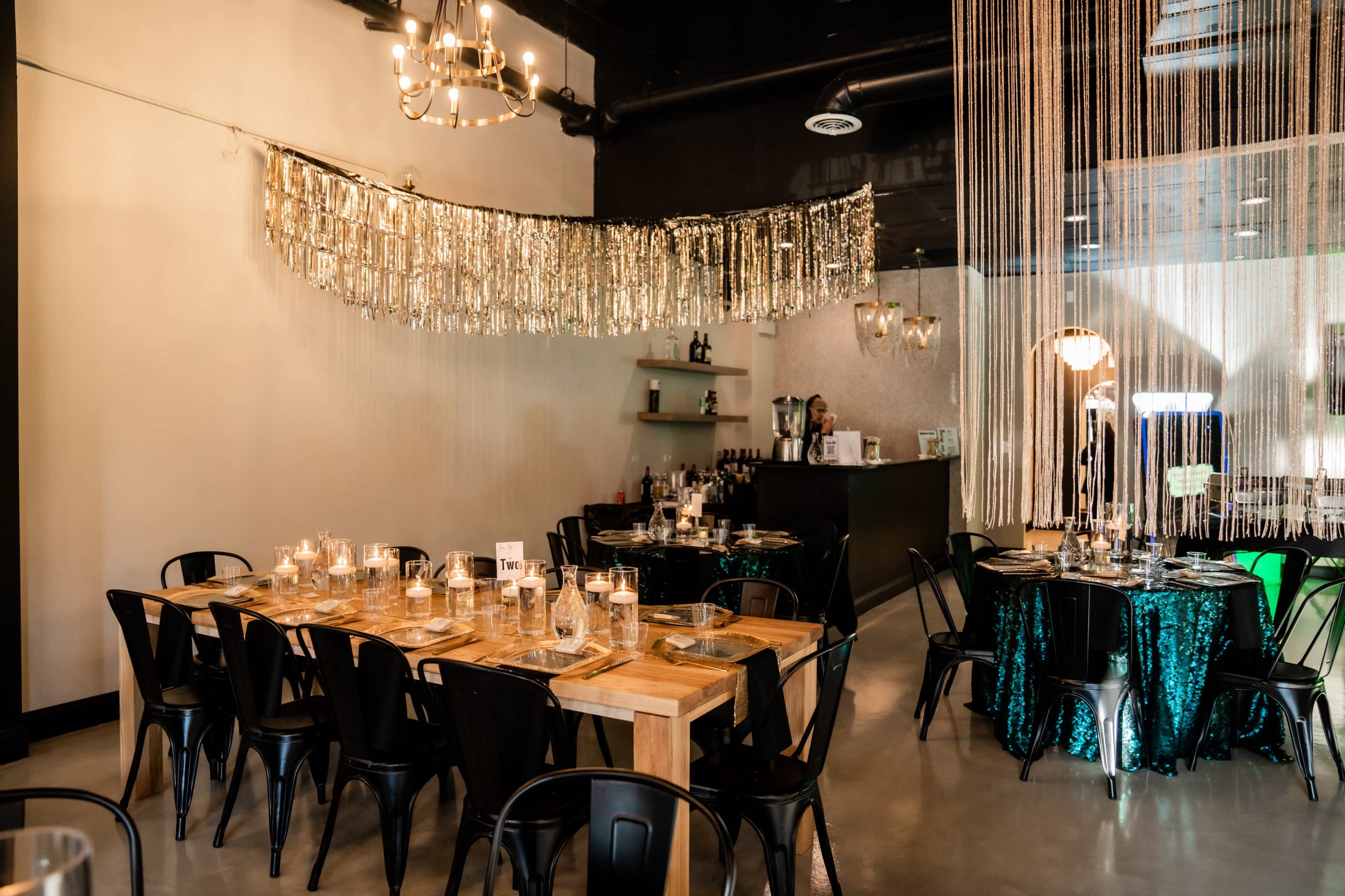 A dining area features two tables, one rectangular made of wood and one round, surrounded by black chairs, with a backdrop of shimmering decorations and a bar in the background.