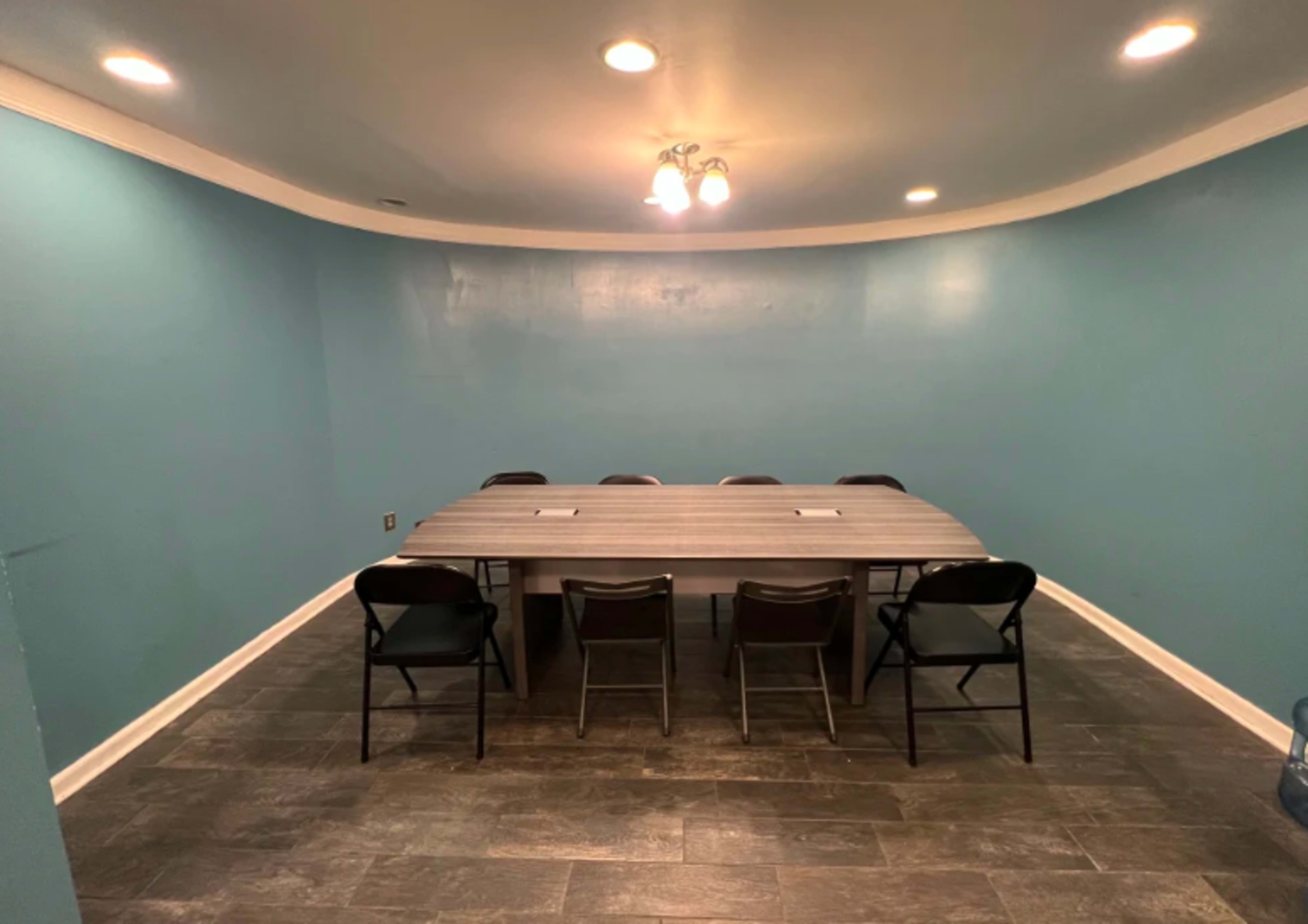 The image shows a spacious room with a rectangular table surrounded by chairs against a blue wall with recessed lighting.