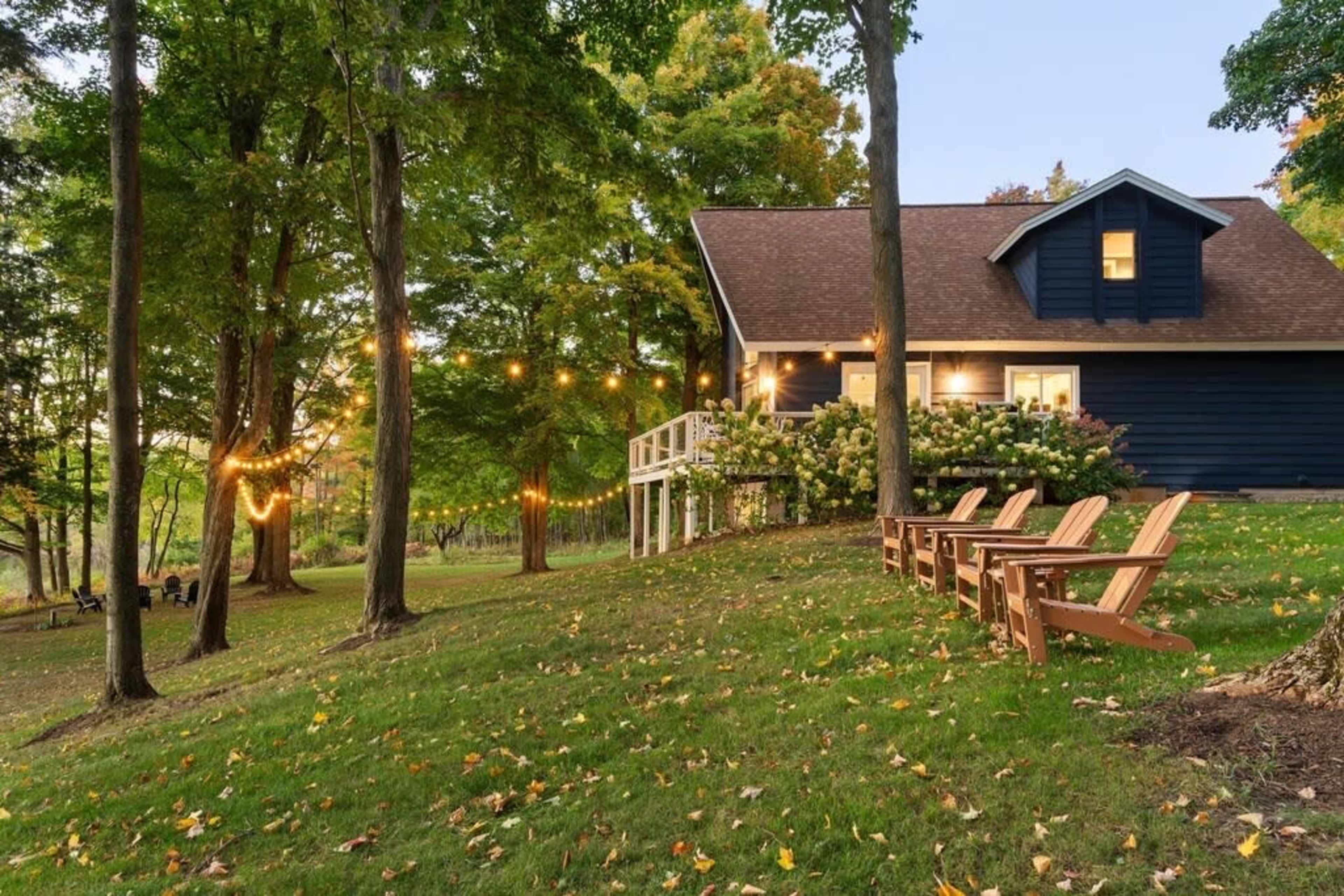 A blue house with a porch is set among trees, adorned with string lights, and features wooden Adirondack chairs on a grassy lawn.