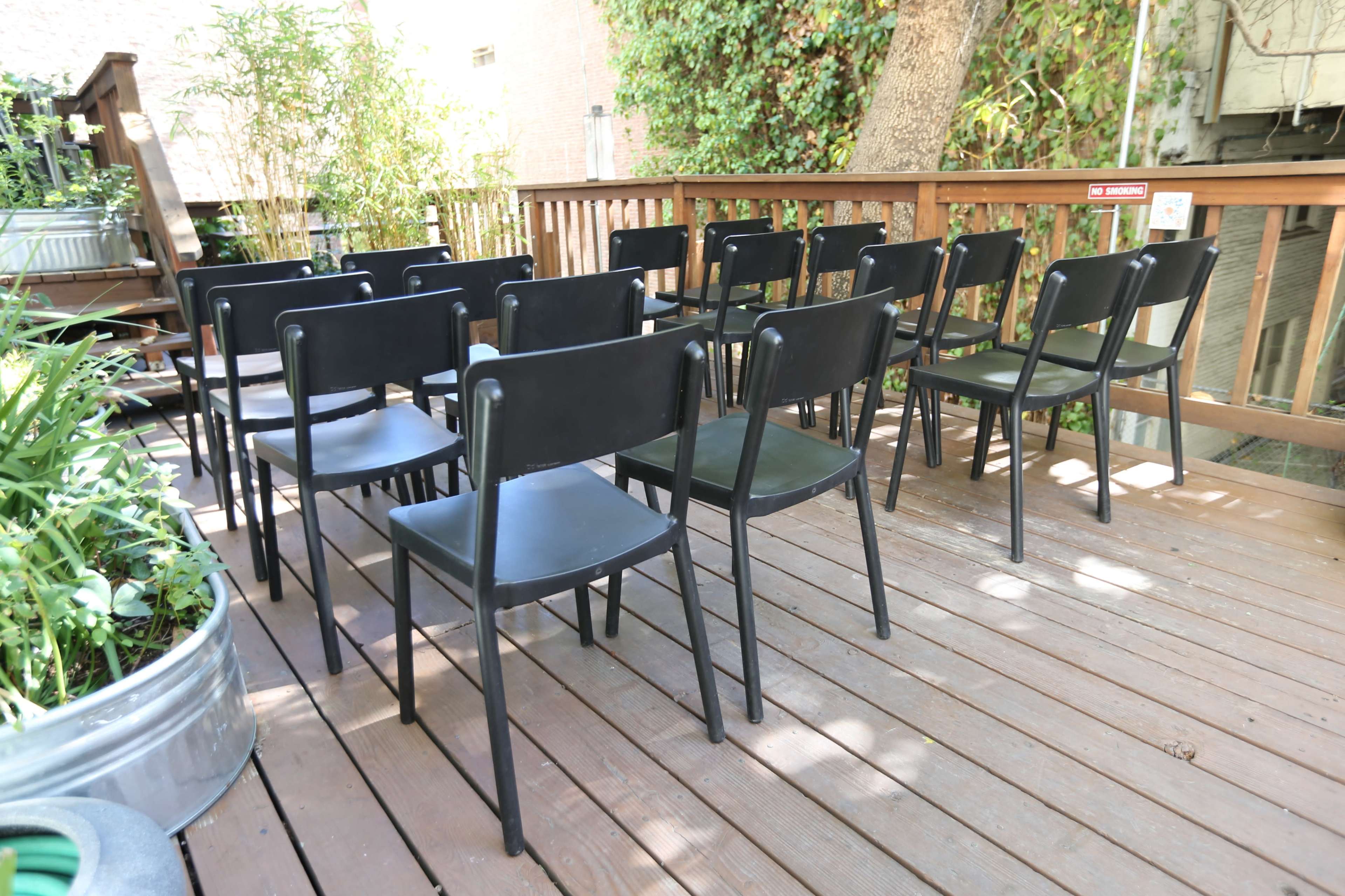 A row of black chairs is set up on a wooden deck surrounded by greenery.