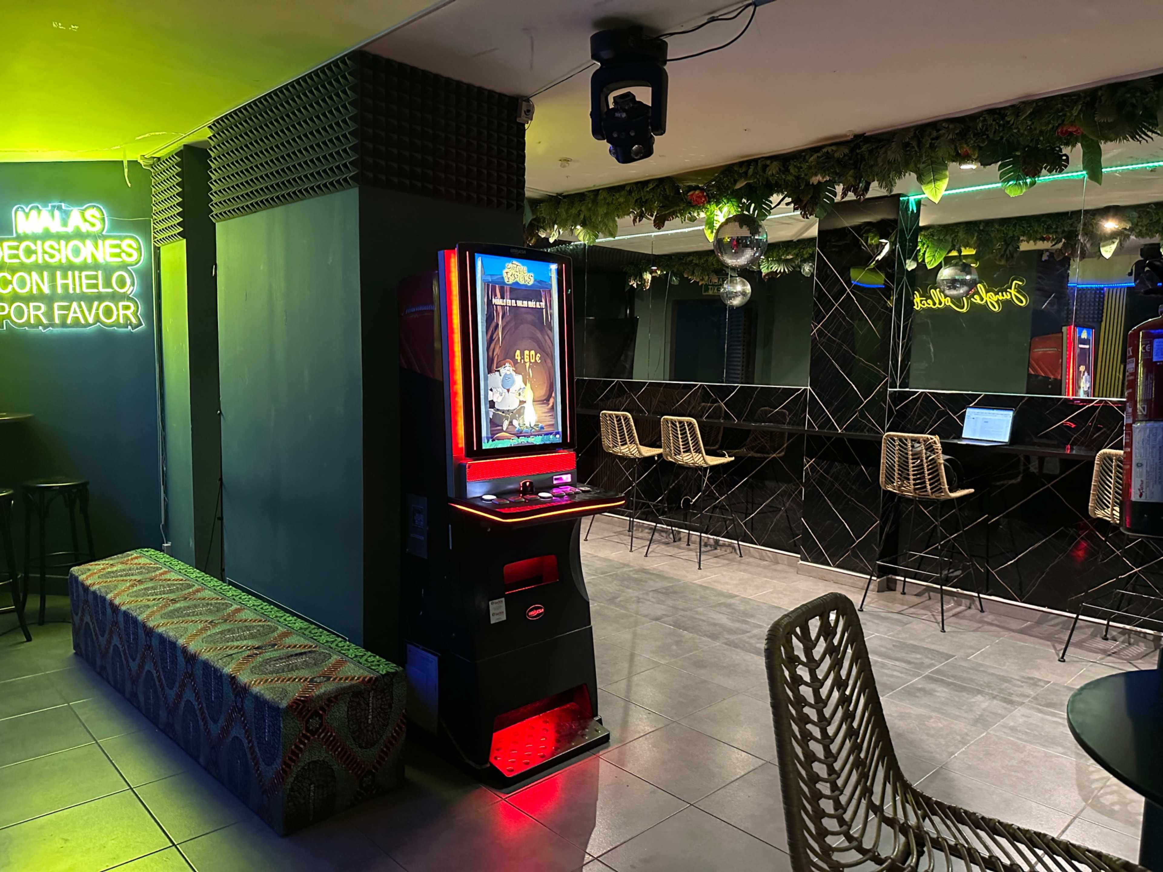 The image shows a dimly lit bar area with a slot machine on the left, a seating area with decorative chairs, and a wall adorned with green foliage and neon signs.