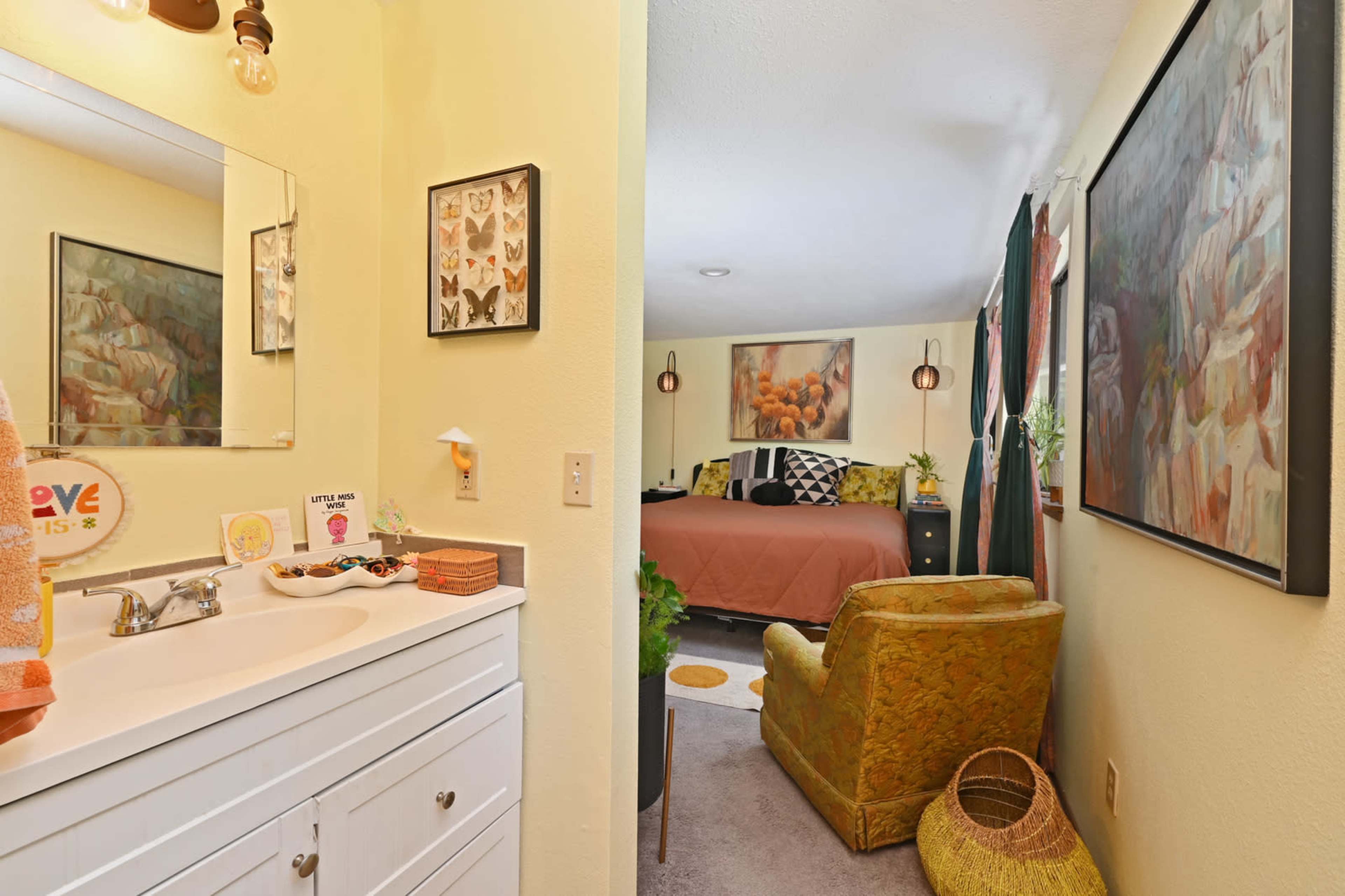 The image shows a bathroom area with a white vanity and a mirror, leading into a cozy bedroom featuring a bed with earthy tones and decorative artwork on the walls.