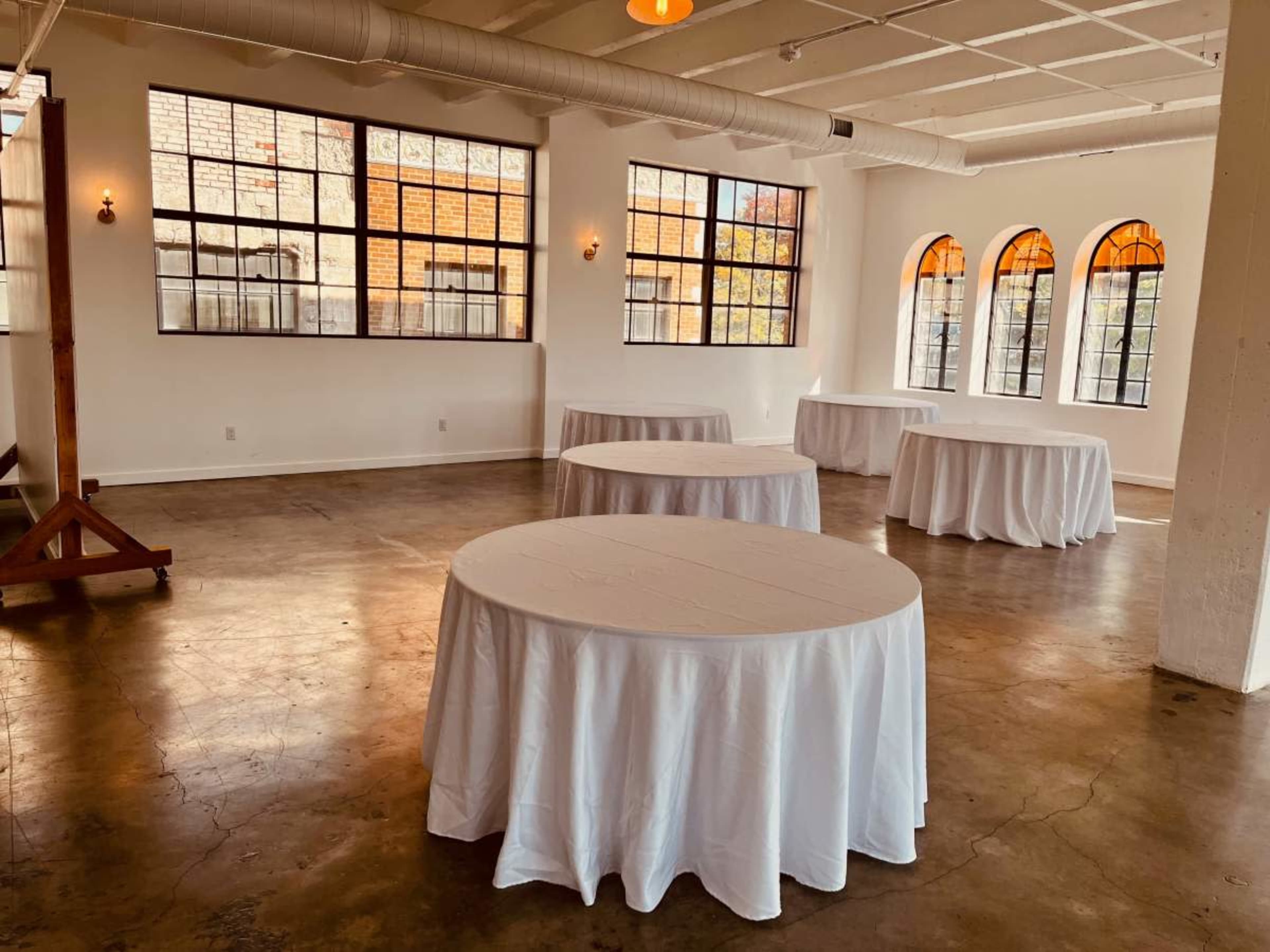 The image shows a spacious room with multiple round tables covered in white tablecloths and large windows allowing natural light to enter.