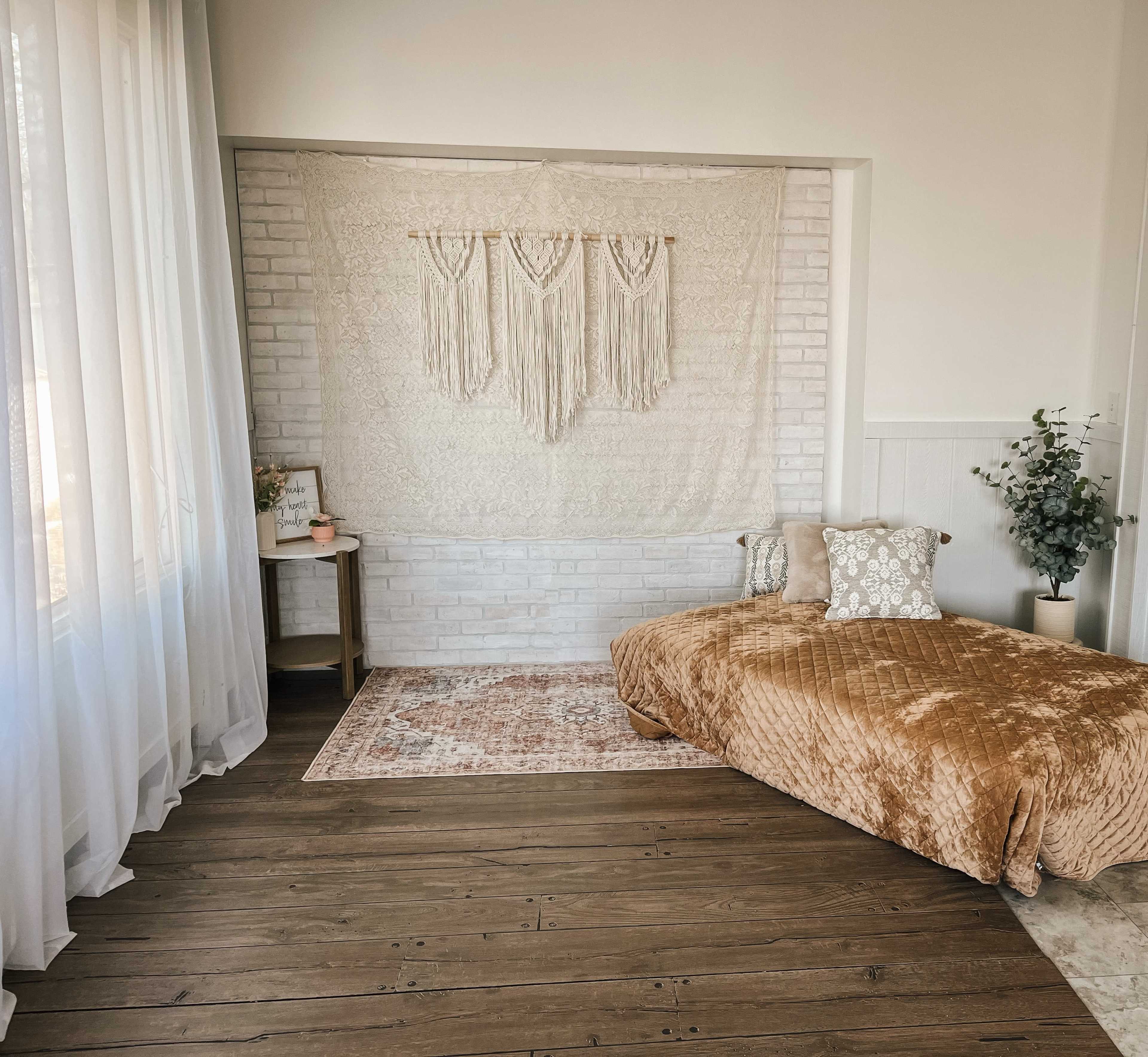 The image shows a bright, minimalist bedroom with a macramé wall hanging, a daybed covered in a textured blanket, and a small side table next to a potted plant.