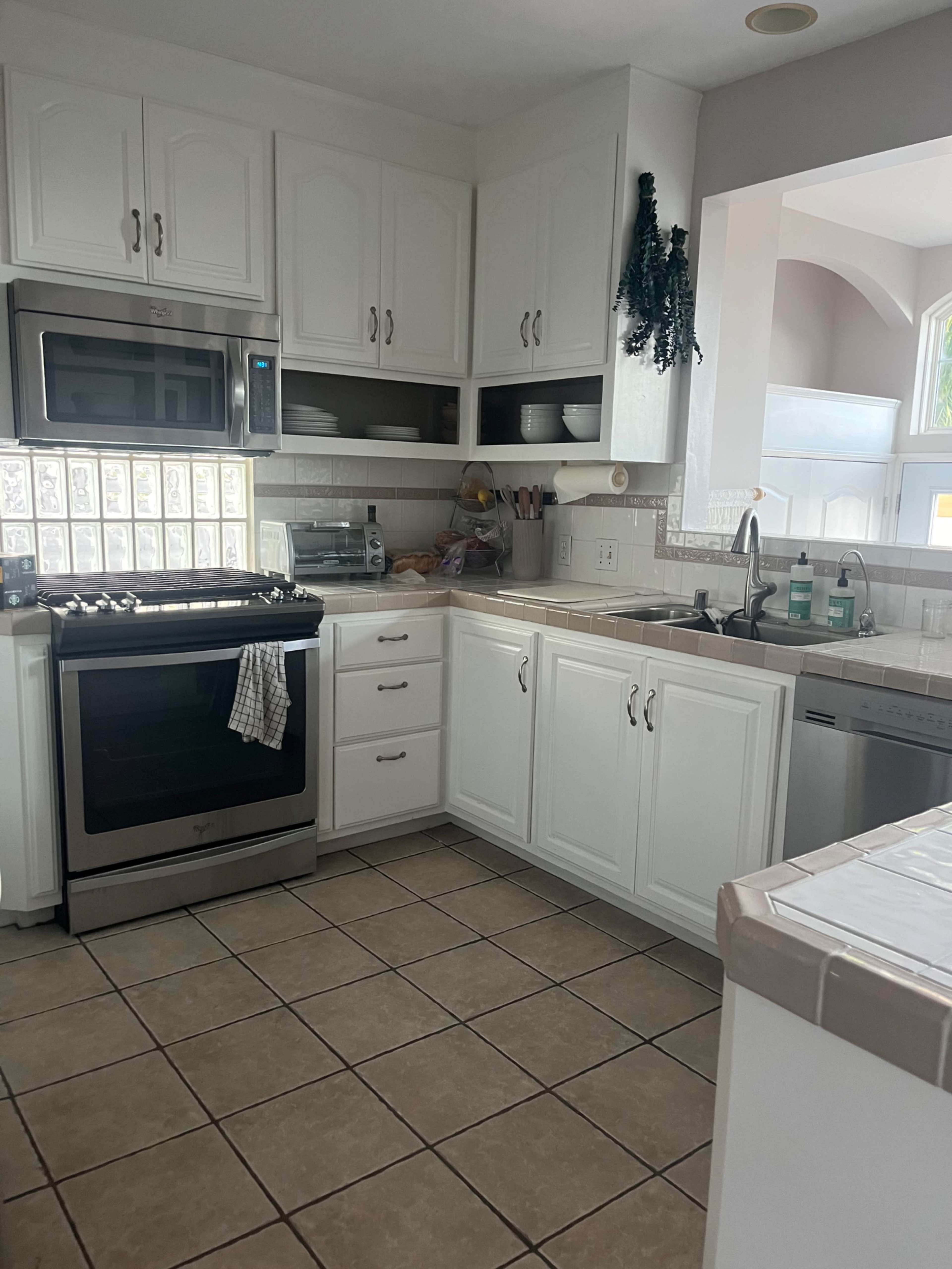 The image shows a kitchen with white cabinets, a stainless steel stove, and a sink, featuring tiled flooring and a variety of kitchen appliances.