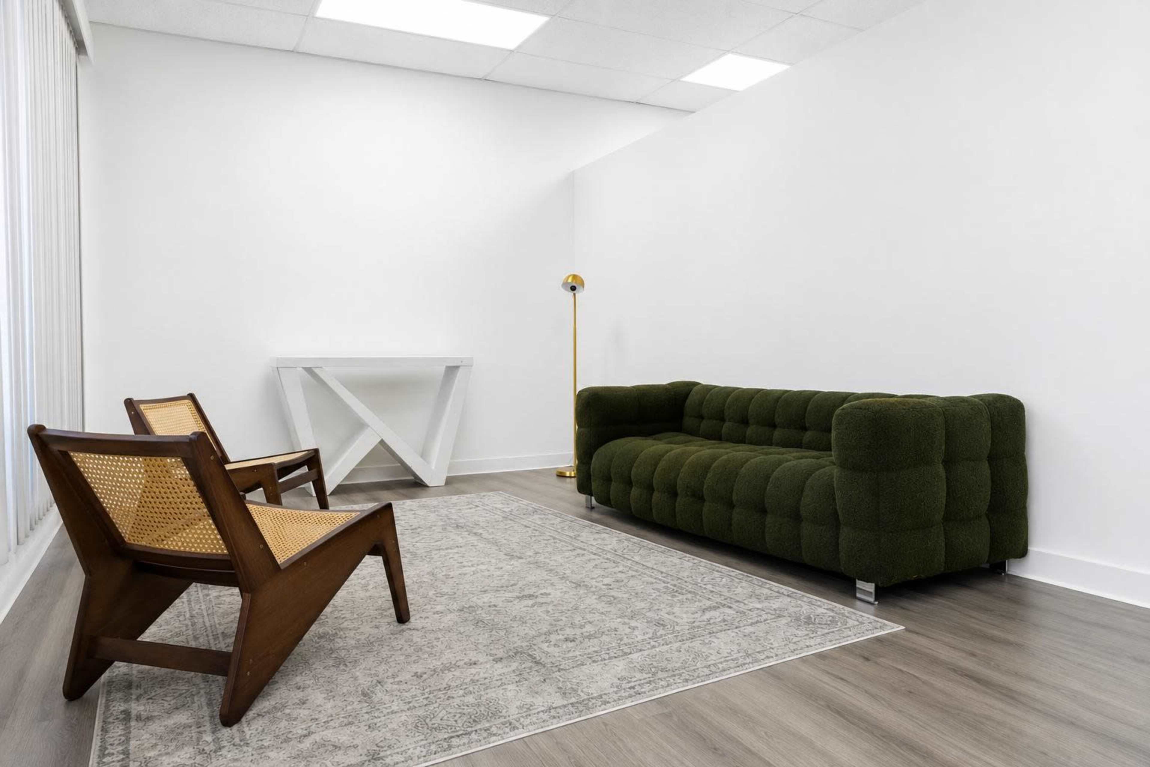 The image shows a modern living space with a green upholstered sofa, two wooden chairs, a white table, and a light grey rug on a hardwood floor.