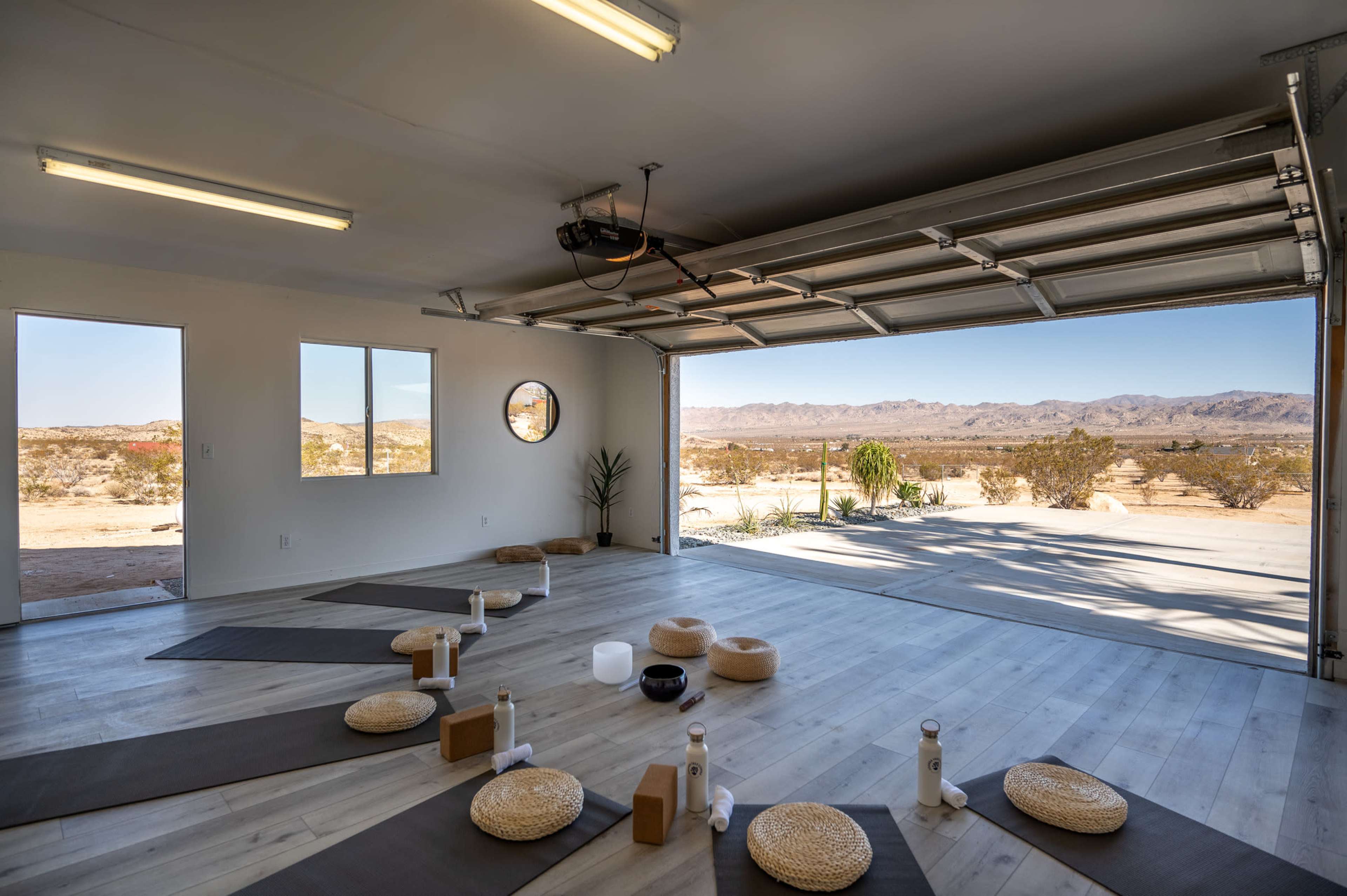 A spacious, minimalist studio is set up for a yoga session, featuring mats and props arranged on the floor, with large garage doors open to reveal a desert landscape.