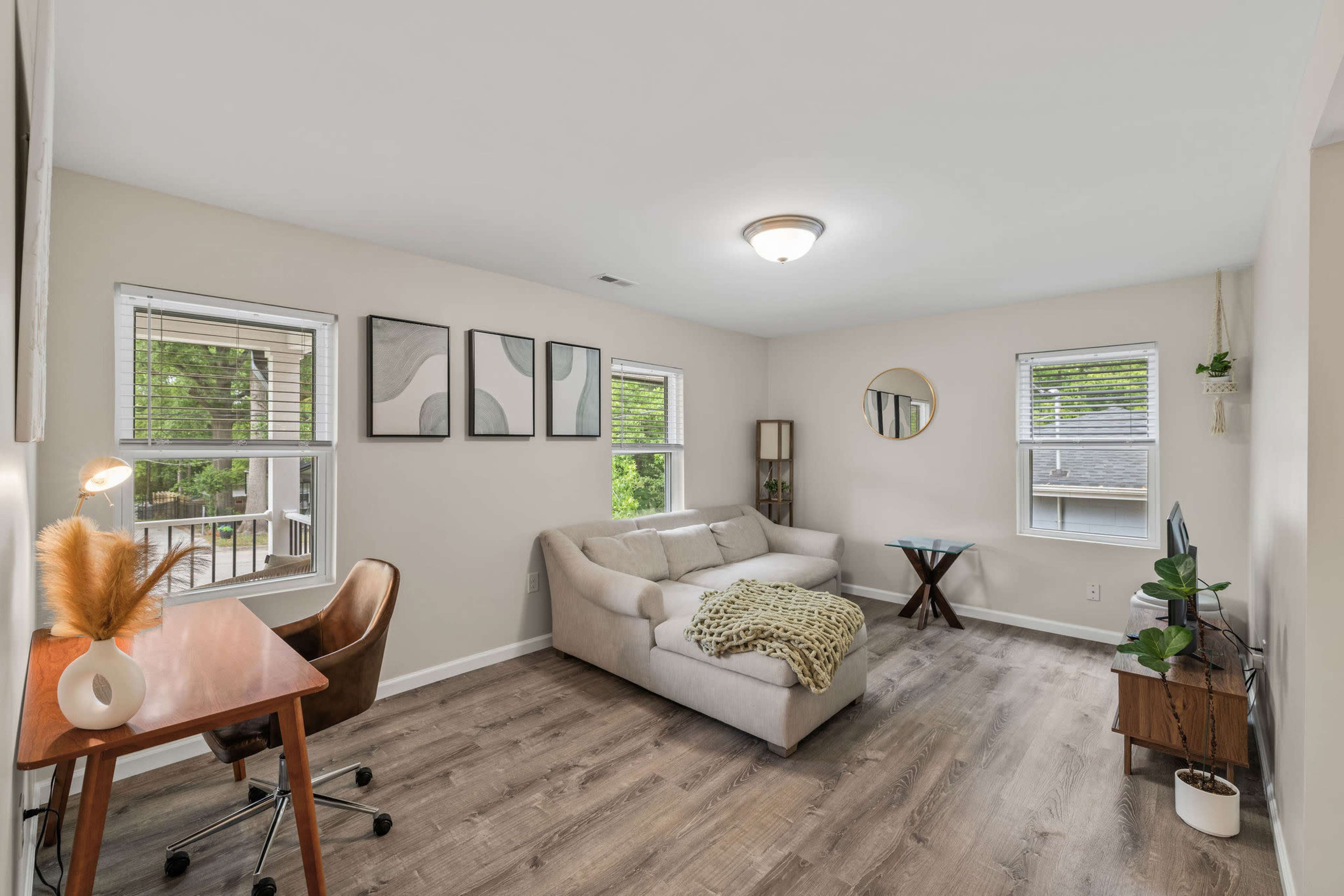 The image shows a light-colored living room with a sofa, a wooden desk, and large windows allowing natural light in.