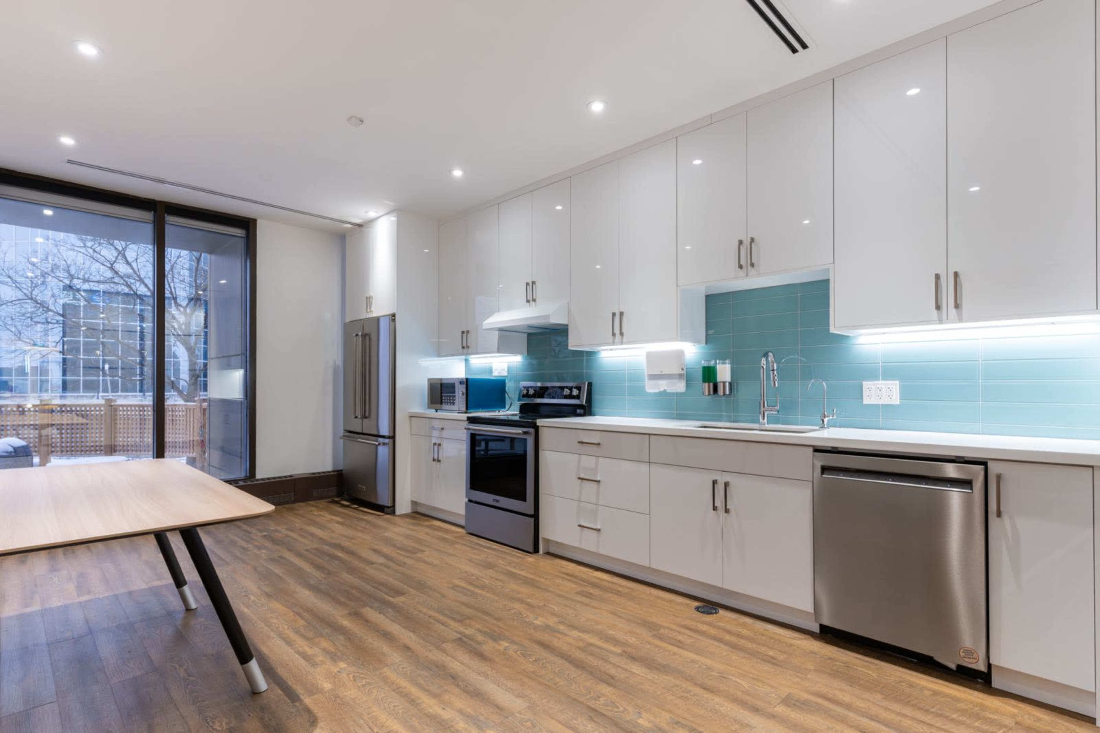 A modern kitchen features white cabinets, stainless steel appliances, and a wooden table, with a large window overlooking an outdoor area.