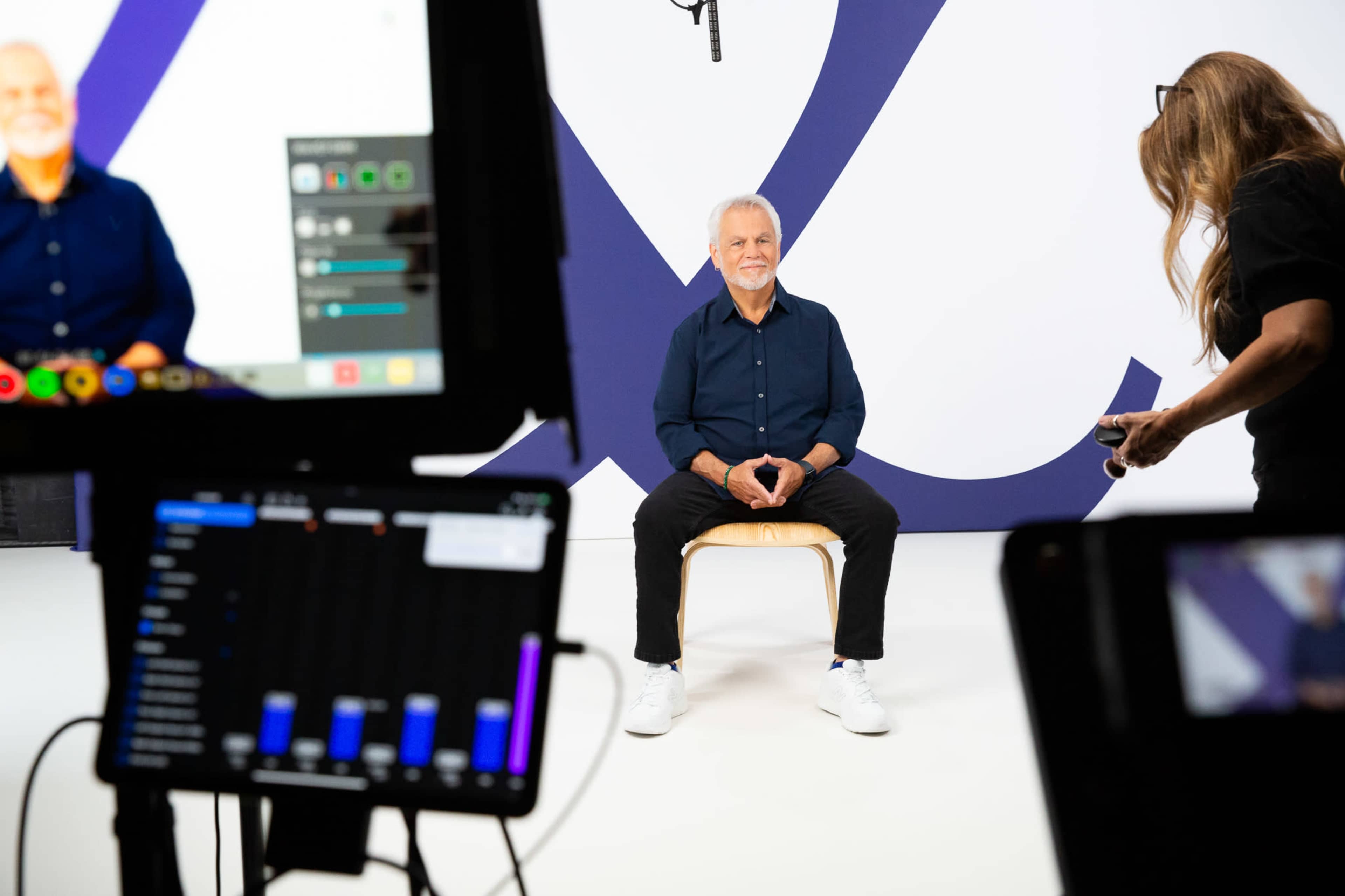 A man sits on a stool in a studio setting while a woman adjusts a camera and lighting equipment nearby.