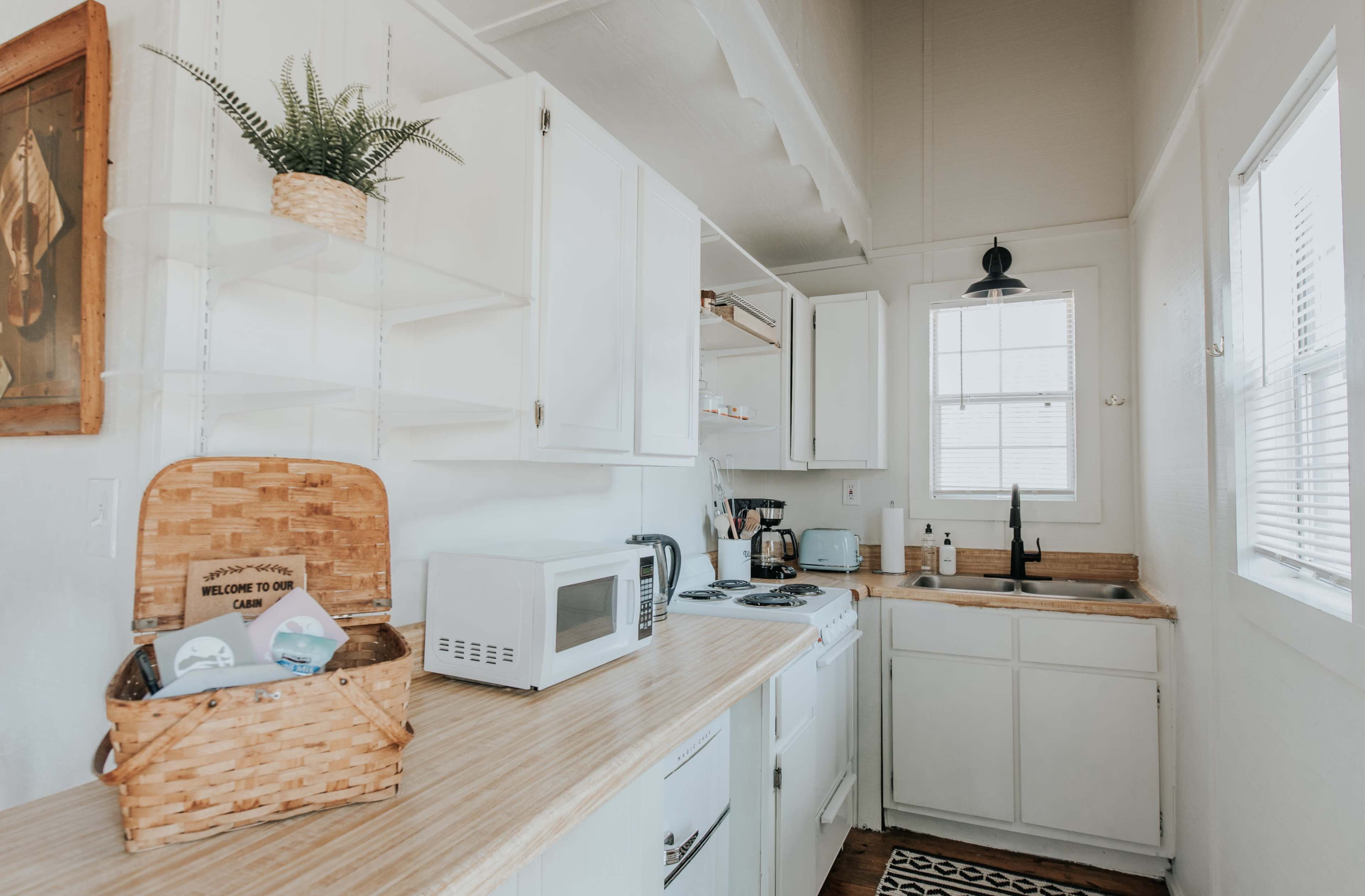 A bright, small kitchen with white cabinets, a microwave, and a wooden countertop lined with a basket and a sink.