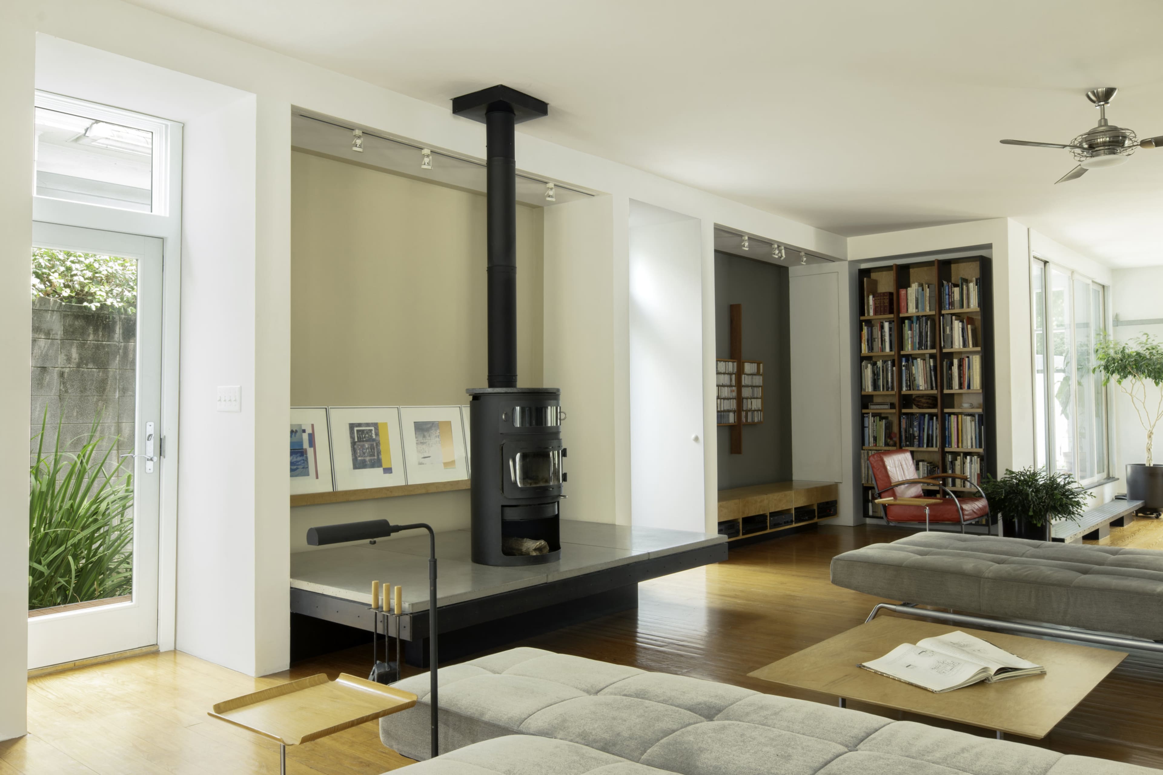 A modern living room features a prominent black wood stove, large windows, and sleek furniture arranged around a hardwood floor.