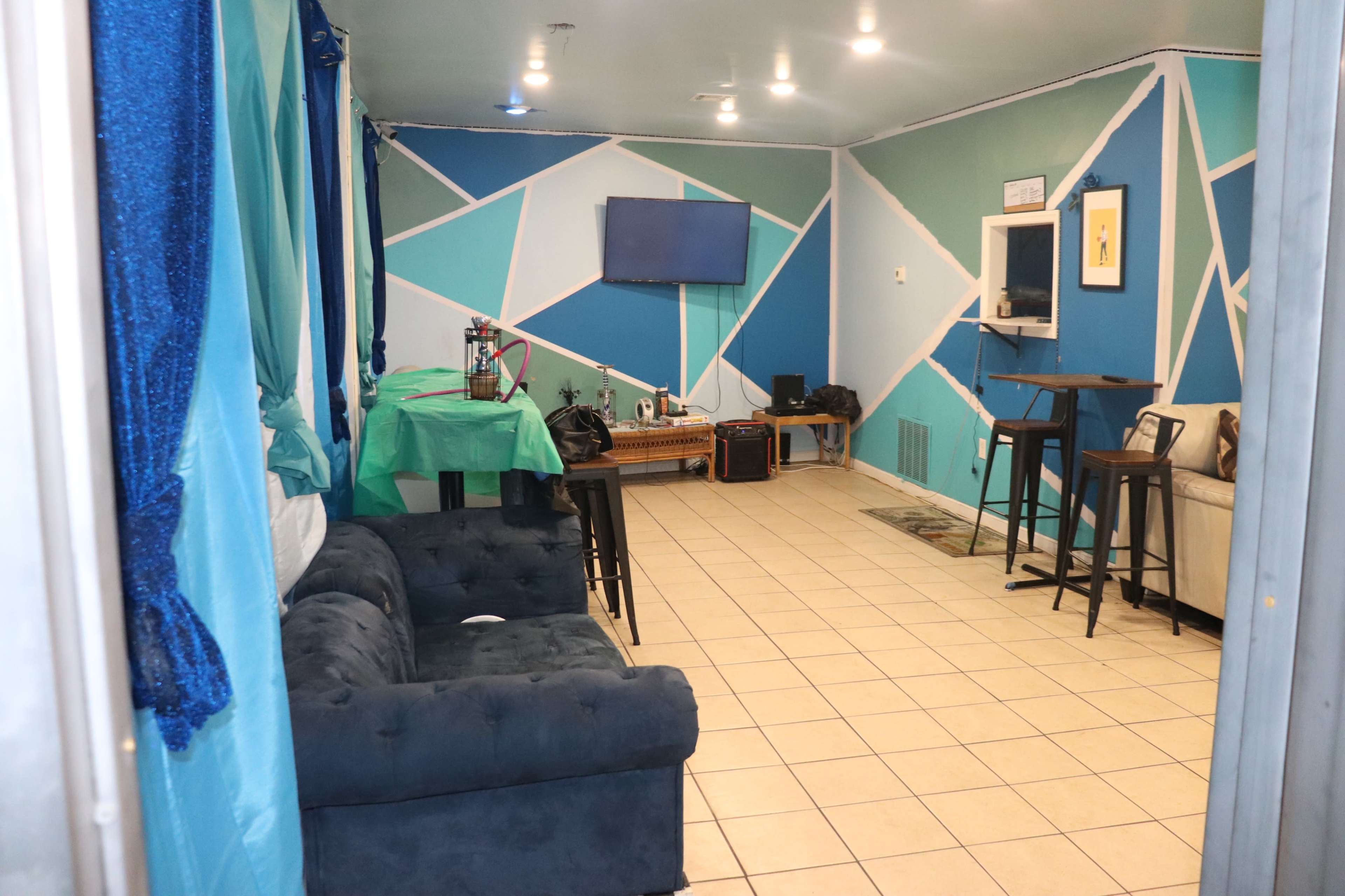 A room with blue and green geometric wall designs, featuring a sofa, a small table with stools, a television mounted on the wall, and various items arranged around the space.