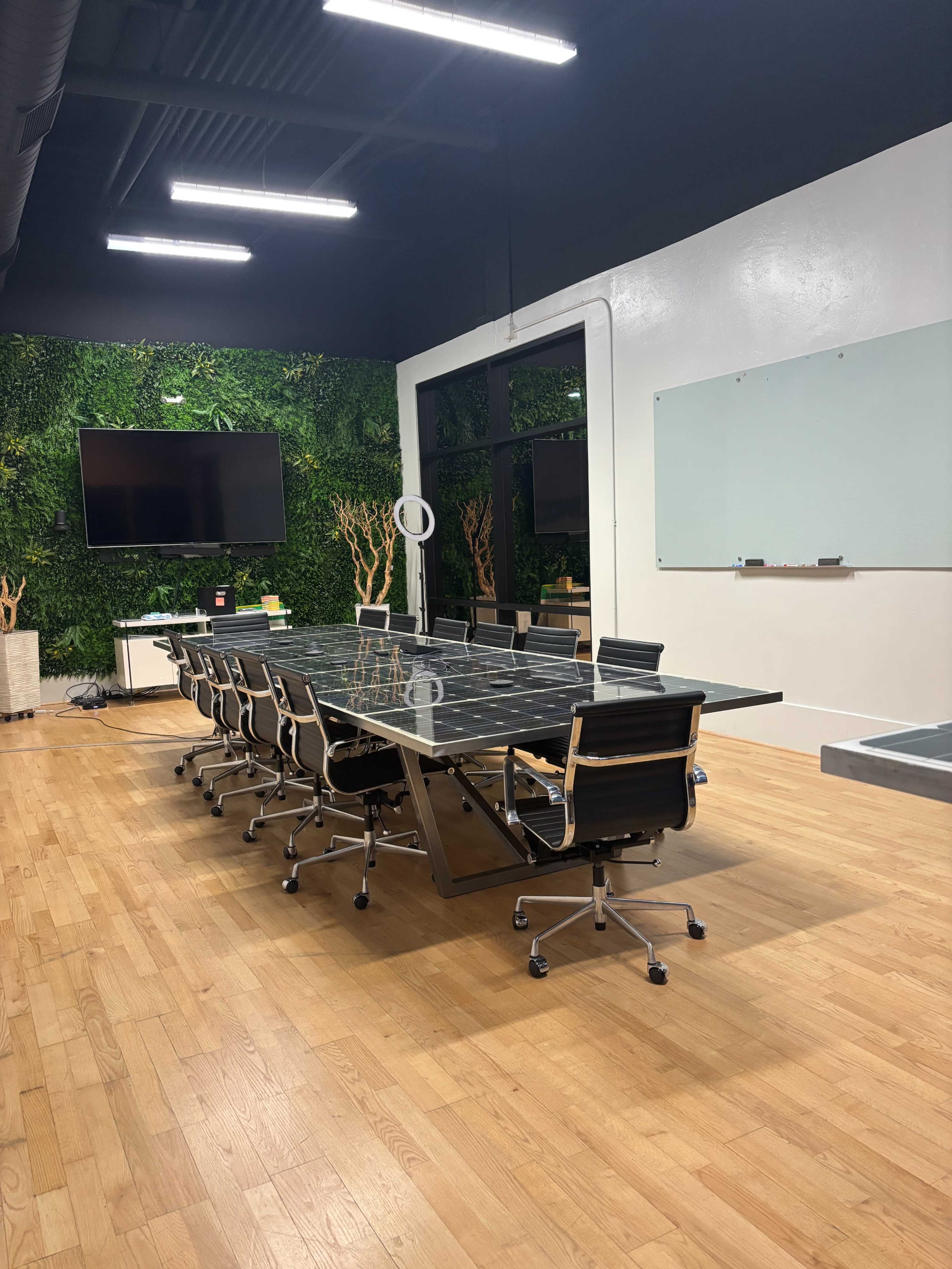 The image shows a modern conference room with a long glass table, black chairs, a large screen on one wall, and greenery accents.