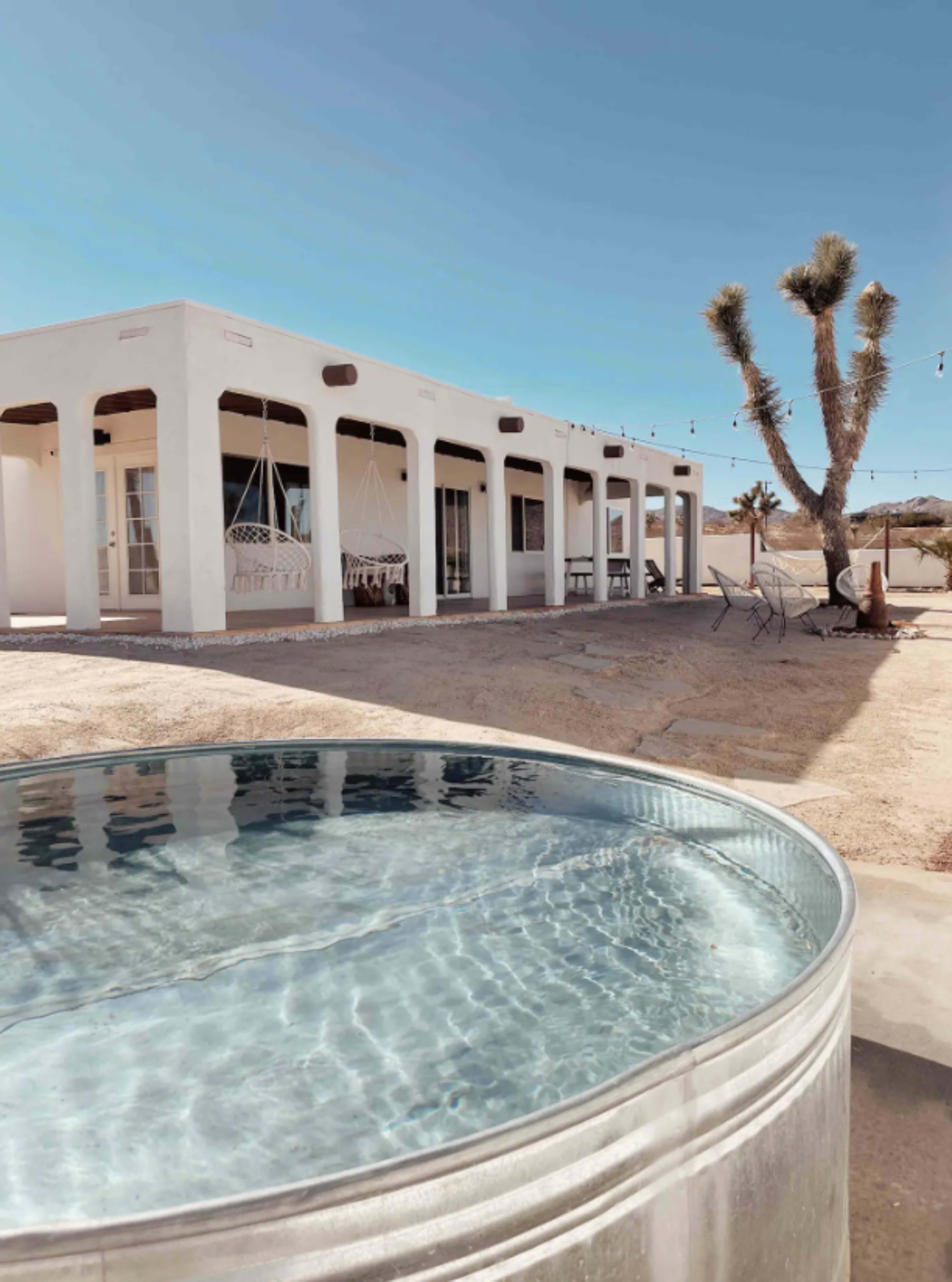 A silver round pool sits in the foreground next to a white adobe-style house with a porch and palm tree in the background.
