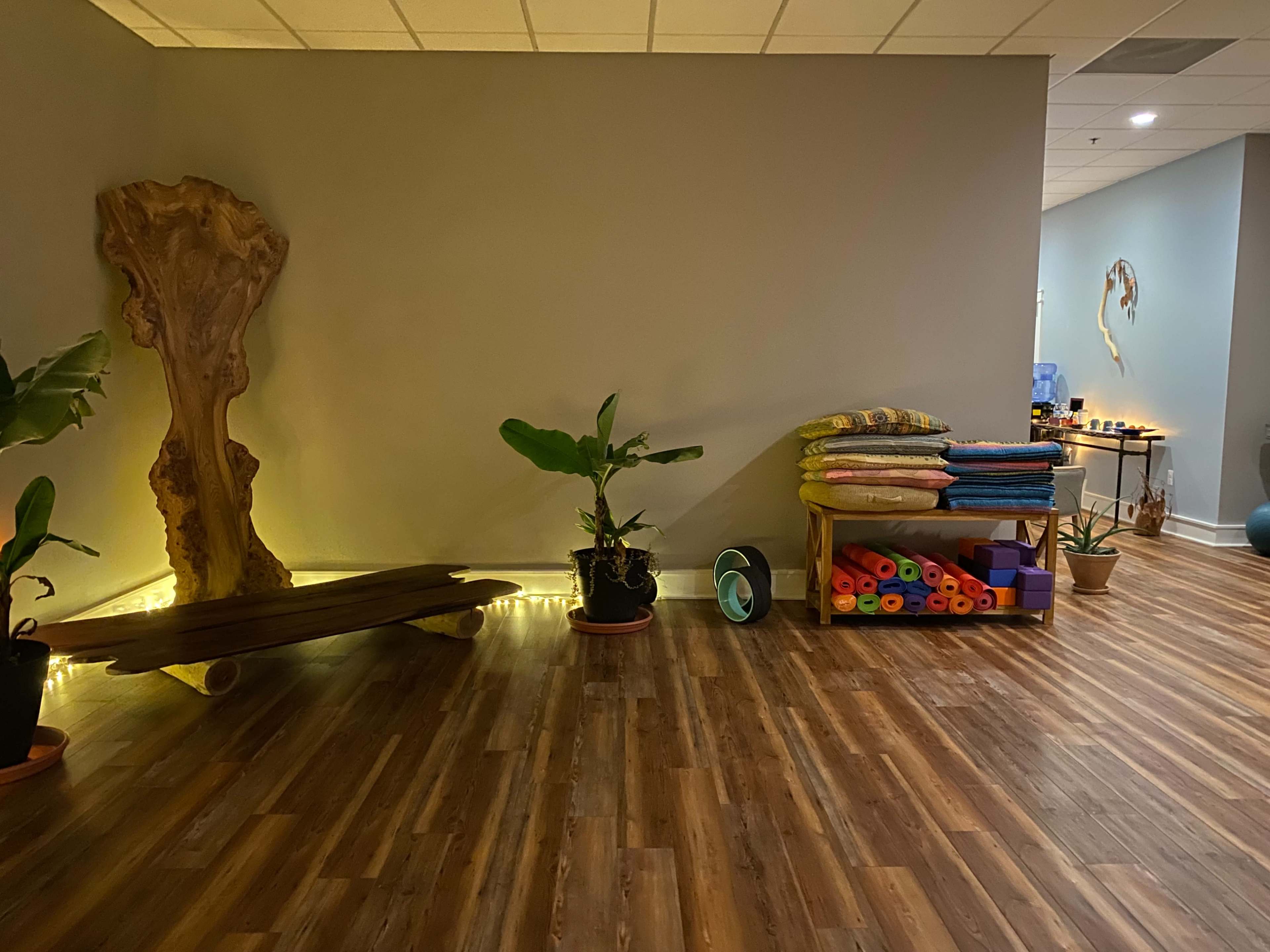 A spacious room with wooden flooring, a decorative wooden structure, plants, and shelves stacked with colorful yoga mats and blankets.