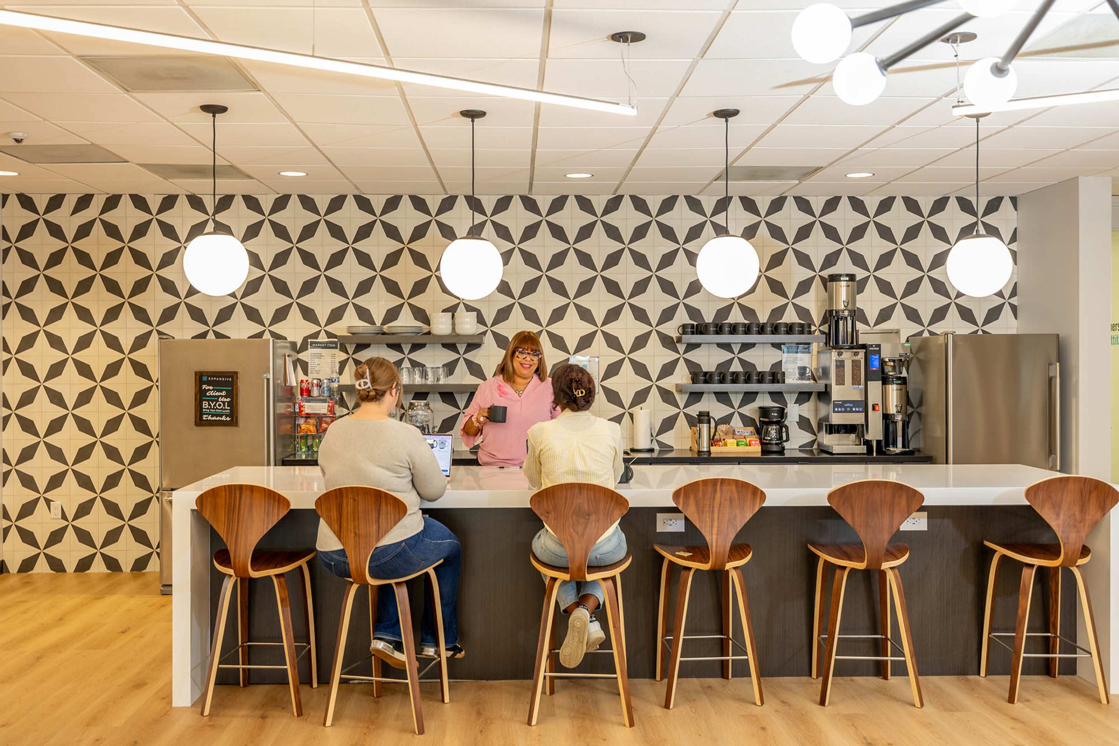 Two people sit at a counter with wooden stools, while a third person stands behind the counter in a modern kitchen featuring geometric patterned wallpaper and pendant lights.