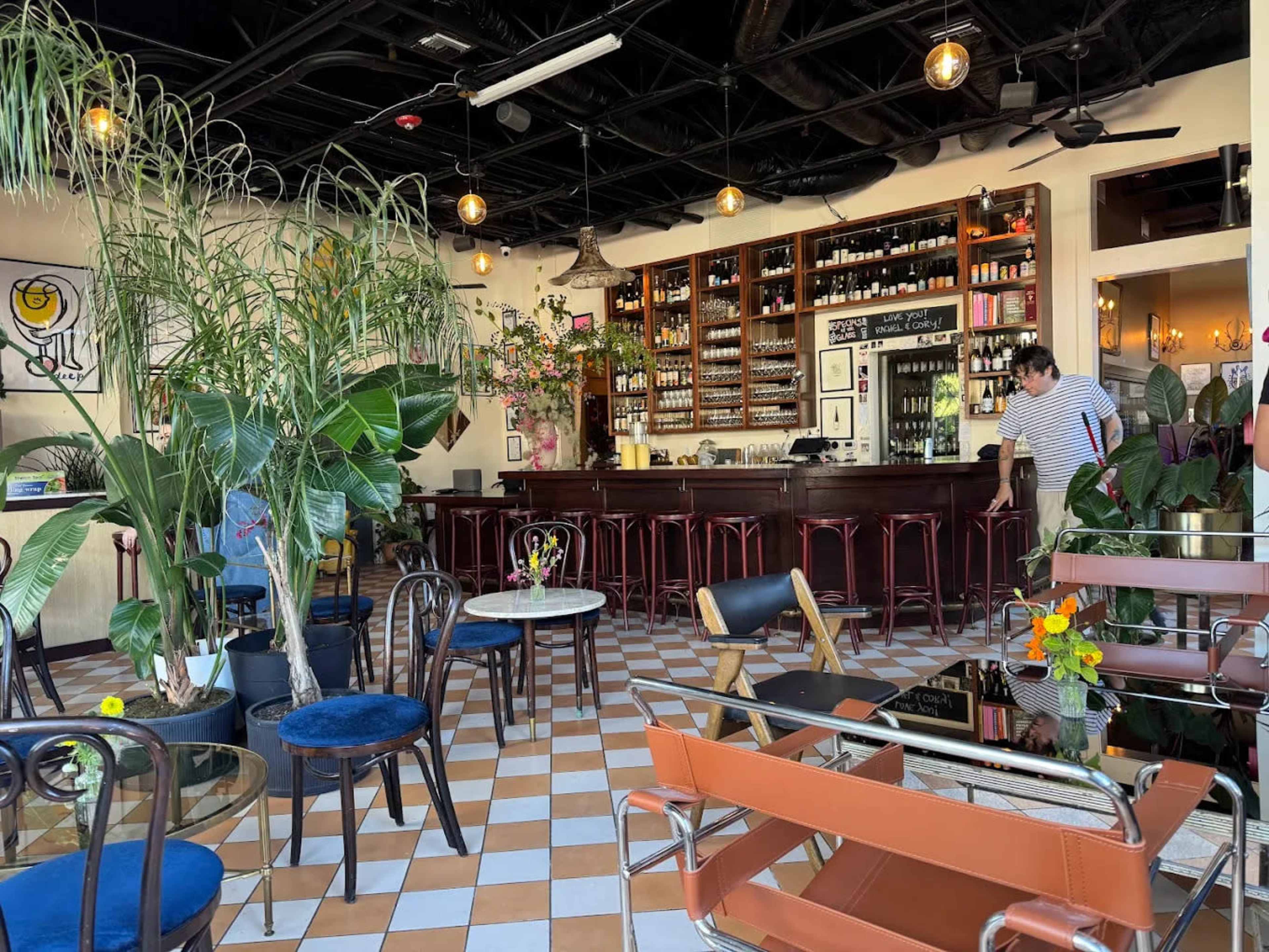 The image shows a stylish café interior featuring a bar, seating arrangements, potted plants, and a checkerboard floor.