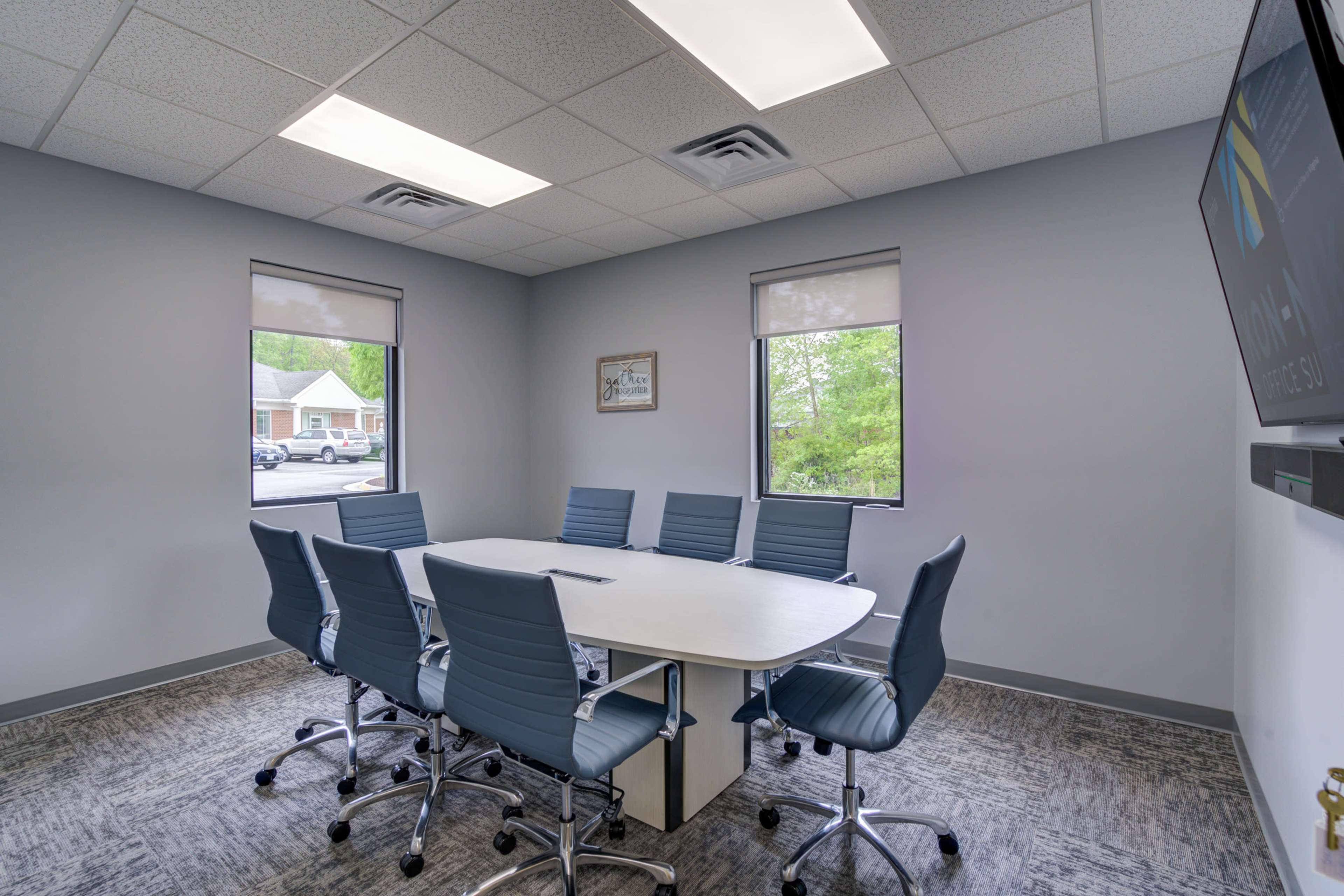 A conference room with a large white table surrounded by eight blue chairs and two windows letting in natural light.