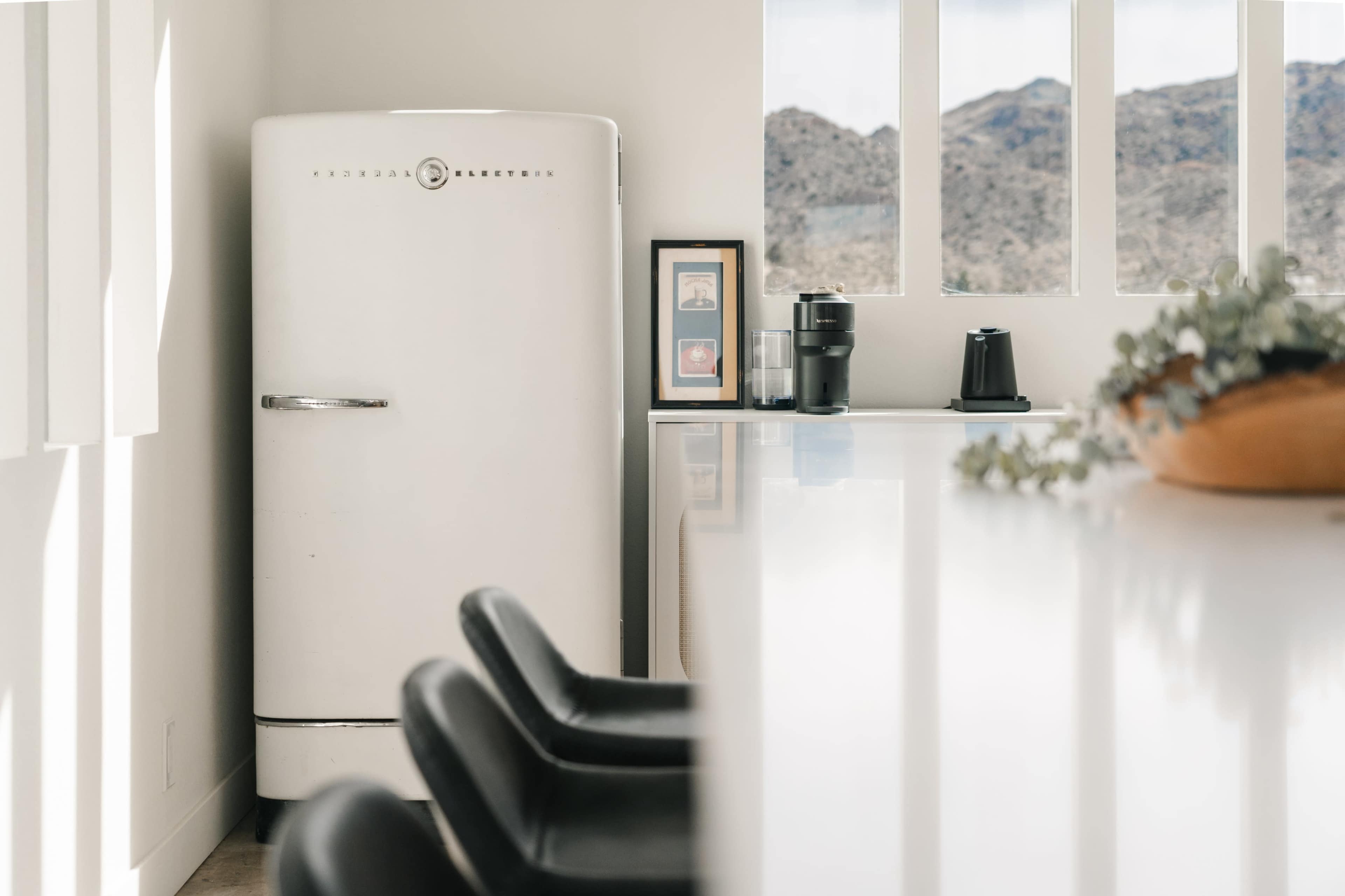 The image shows a white retro refrigerator next to a coffee maker on a countertop with a view of a mountainous landscape through large windows.