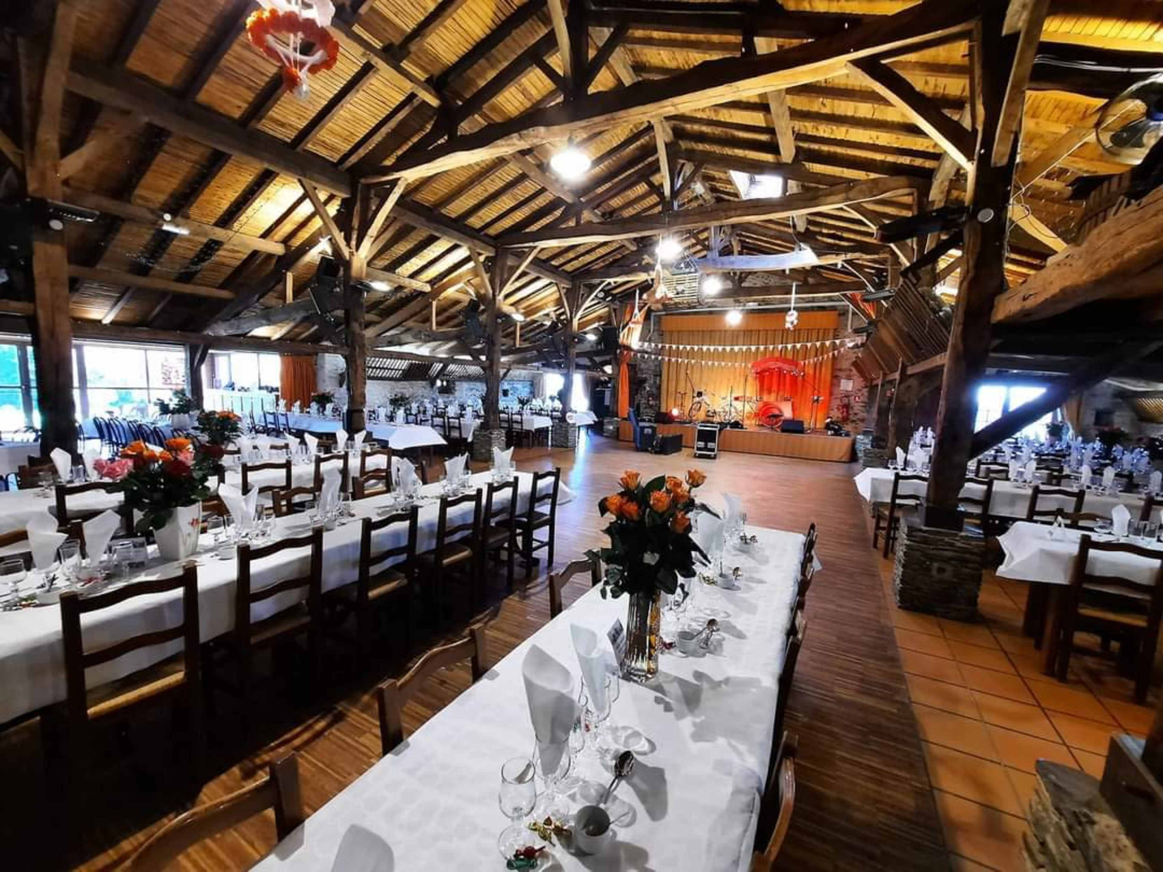 A spacious rustic venue is set with long dining tables adorned with floral centerpieces, complemented by wooden beams and a small stage at the front.