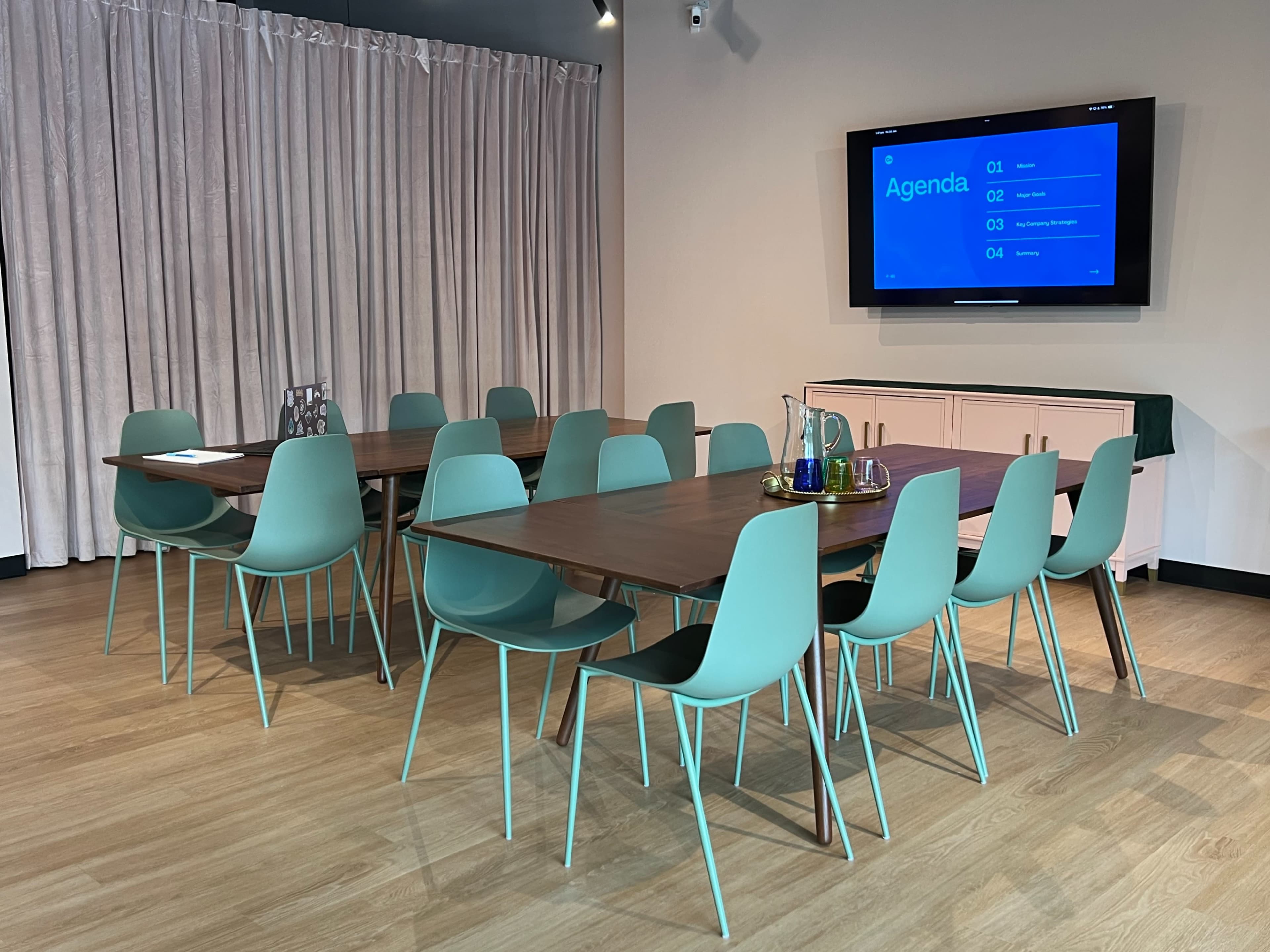A modern meeting room with a large table surrounded by mint green chairs, featuring a screen displaying an agenda and curtains in the background.