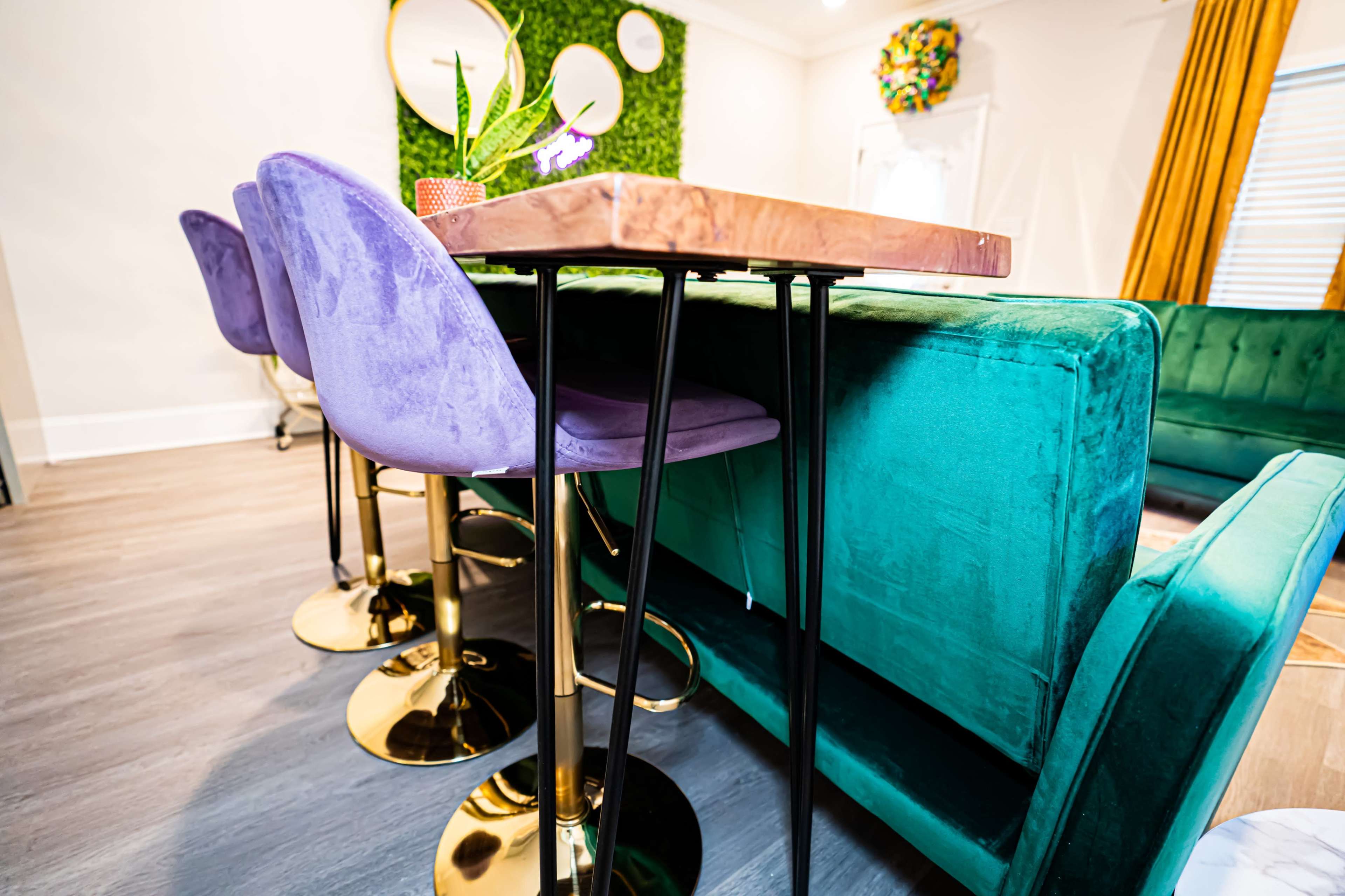 The image shows a modern dining area featuring a tall table with a metallic top, surrounded by purple velvet stools and a green upholstered bench against the wall.