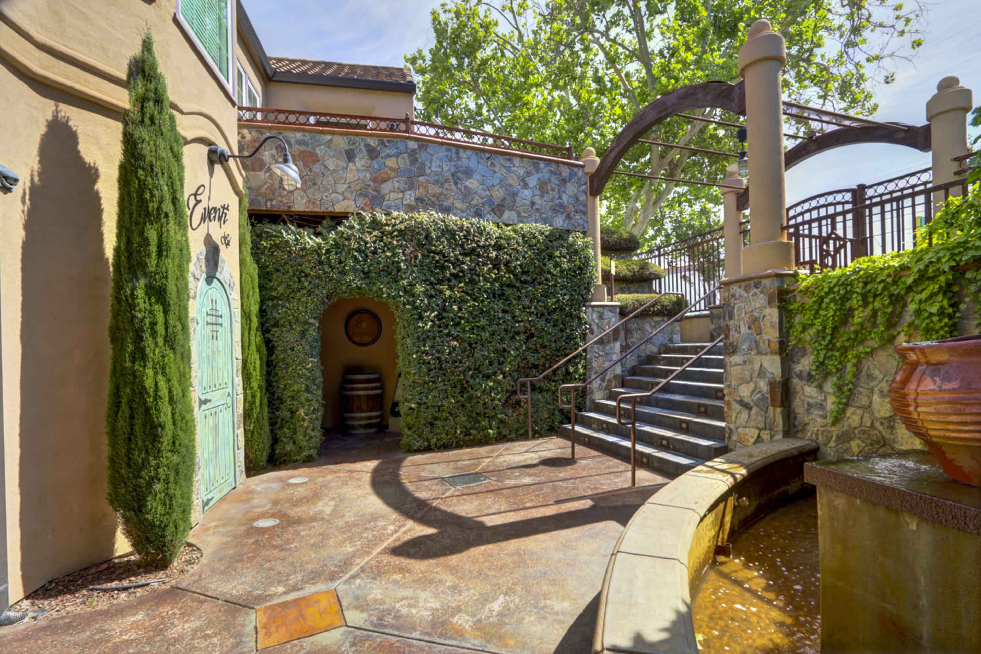A courtyard with a stone fountain, lush greenery, stairs leading up to a patio, and a small entrance marked by a wooden door.