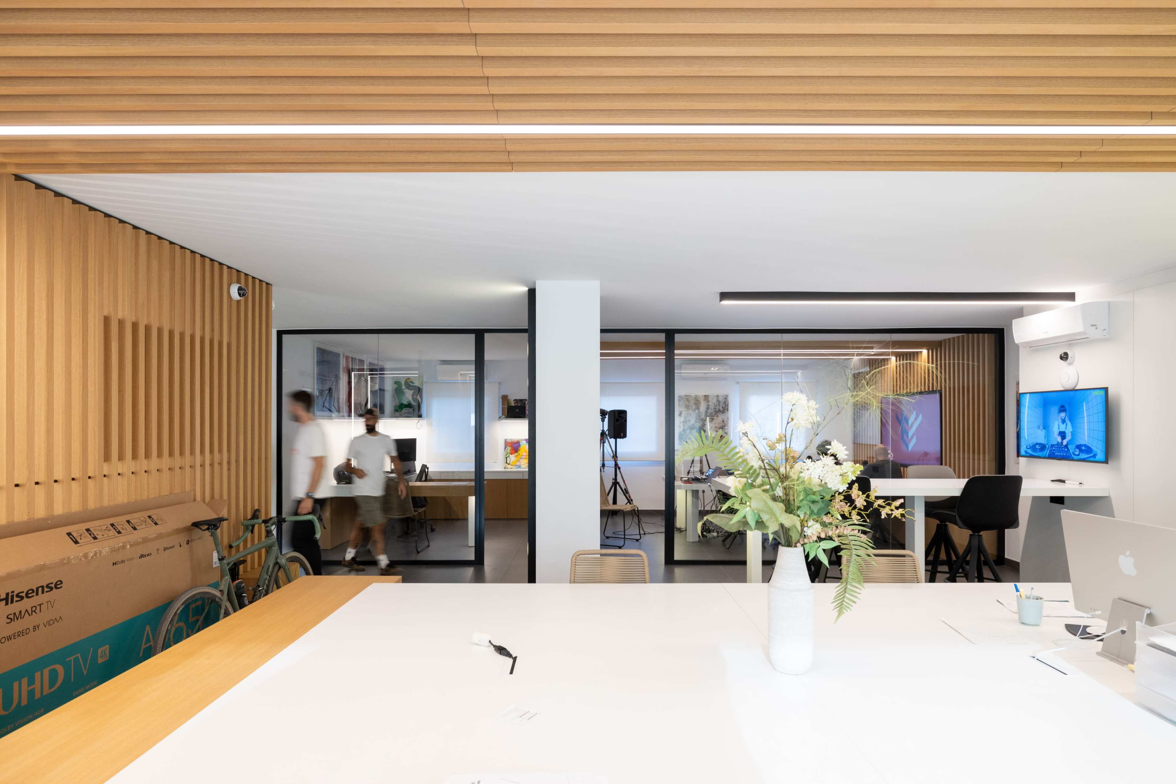 A modern office space featuring wooden paneling, a large white table, and glass walls separating work areas, with people moving around and decorative plants on the table.