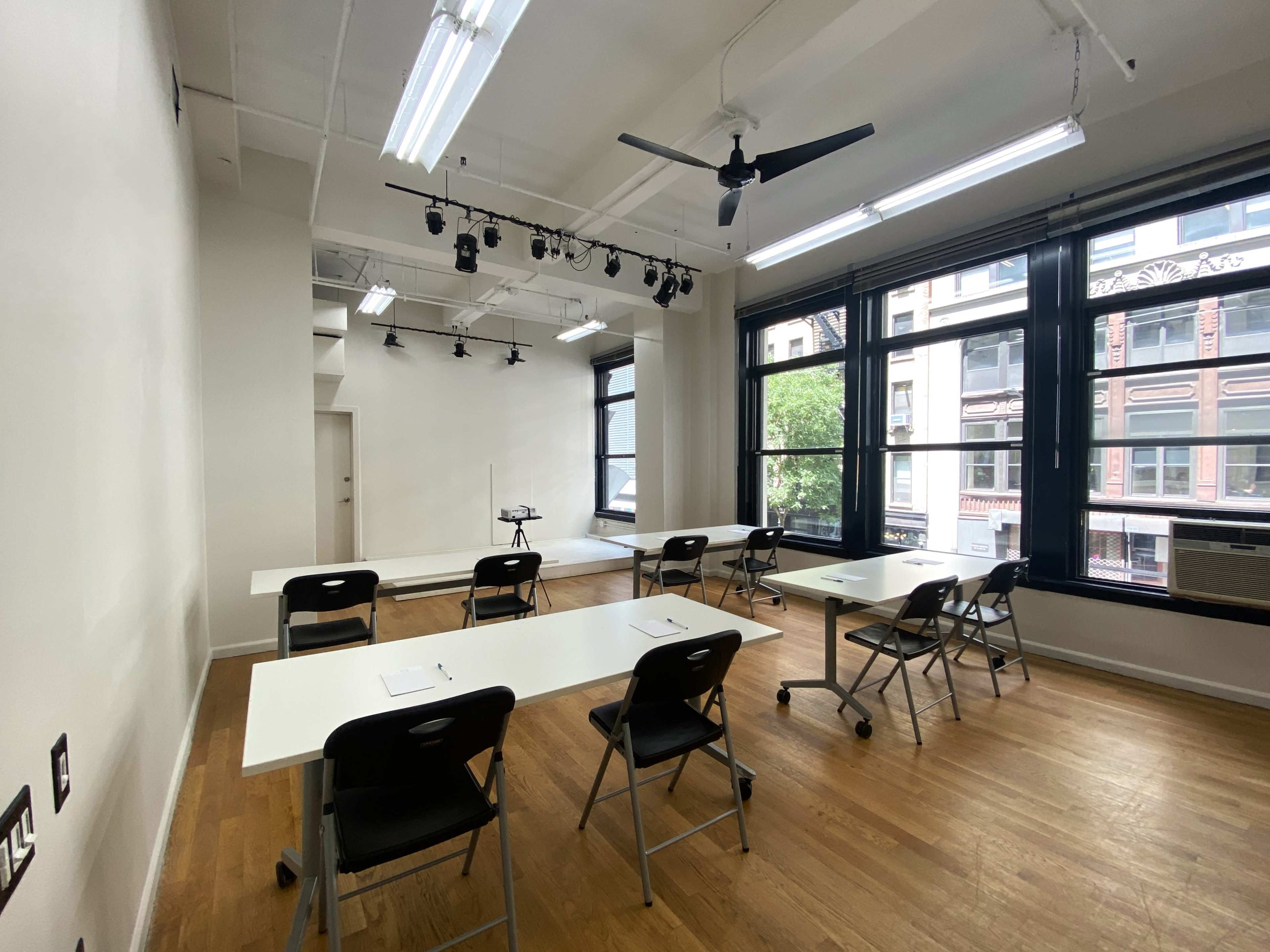The image shows a bright classroom with white tables, black chairs, and large windows overlooking a city street.