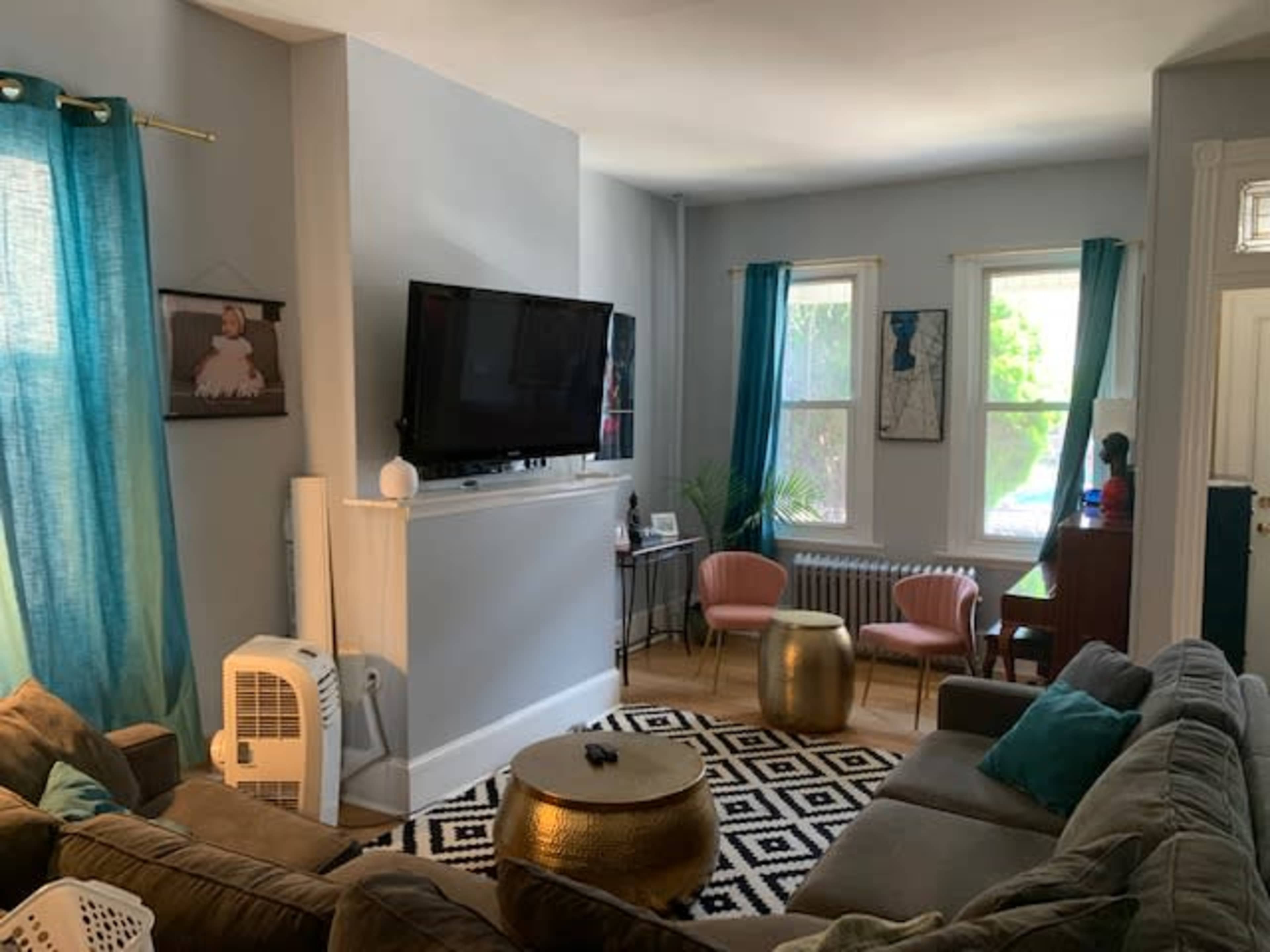 A living room features a sofa, a television mounted on the wall, and two chairs with a coffee table in the center, surrounded by windows with sheer curtains.
