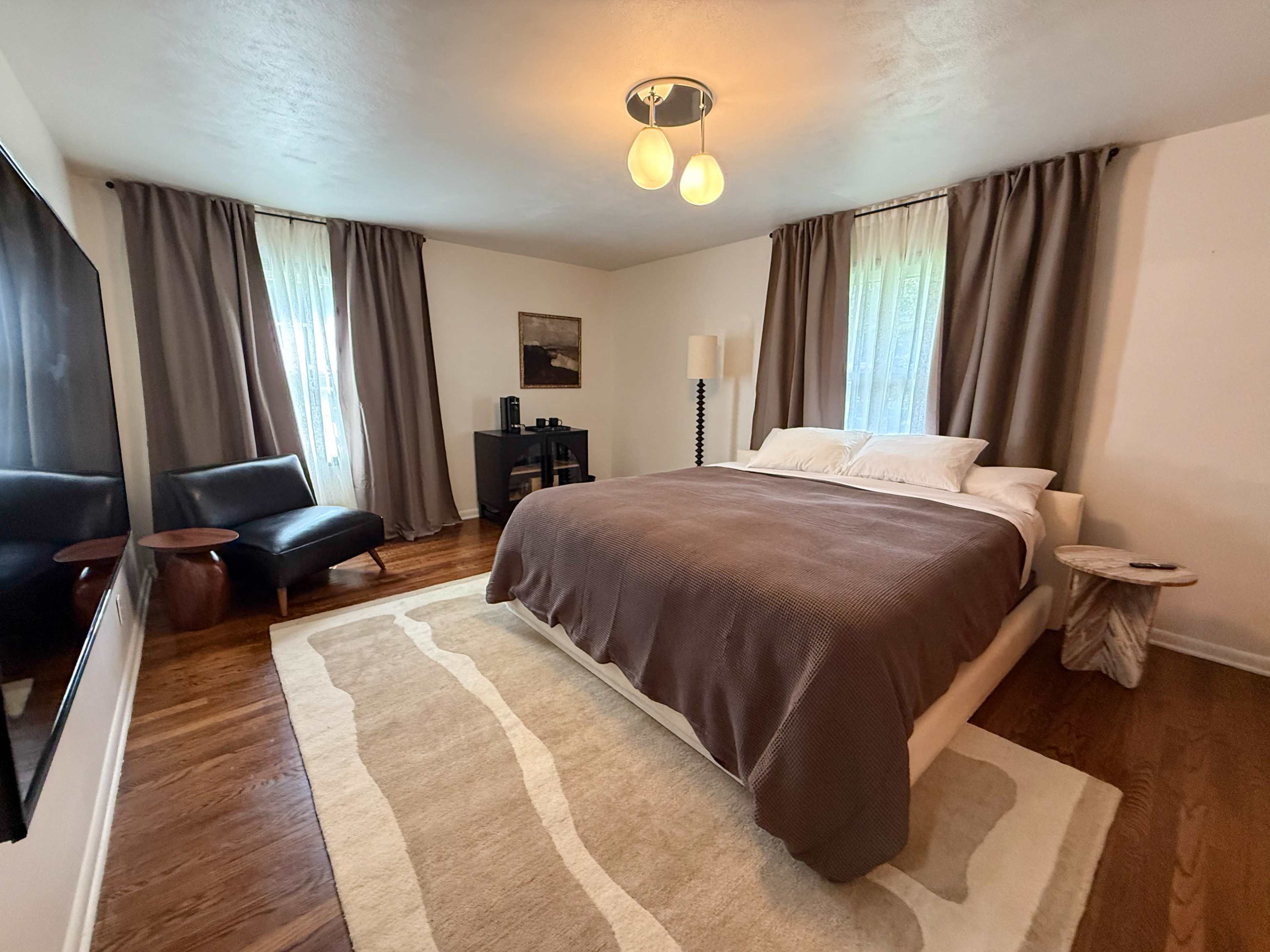 The image shows a modern bedroom featuring a large bed with brown bedding, a black leather chair, a small side table, and a television mounted on the wall.