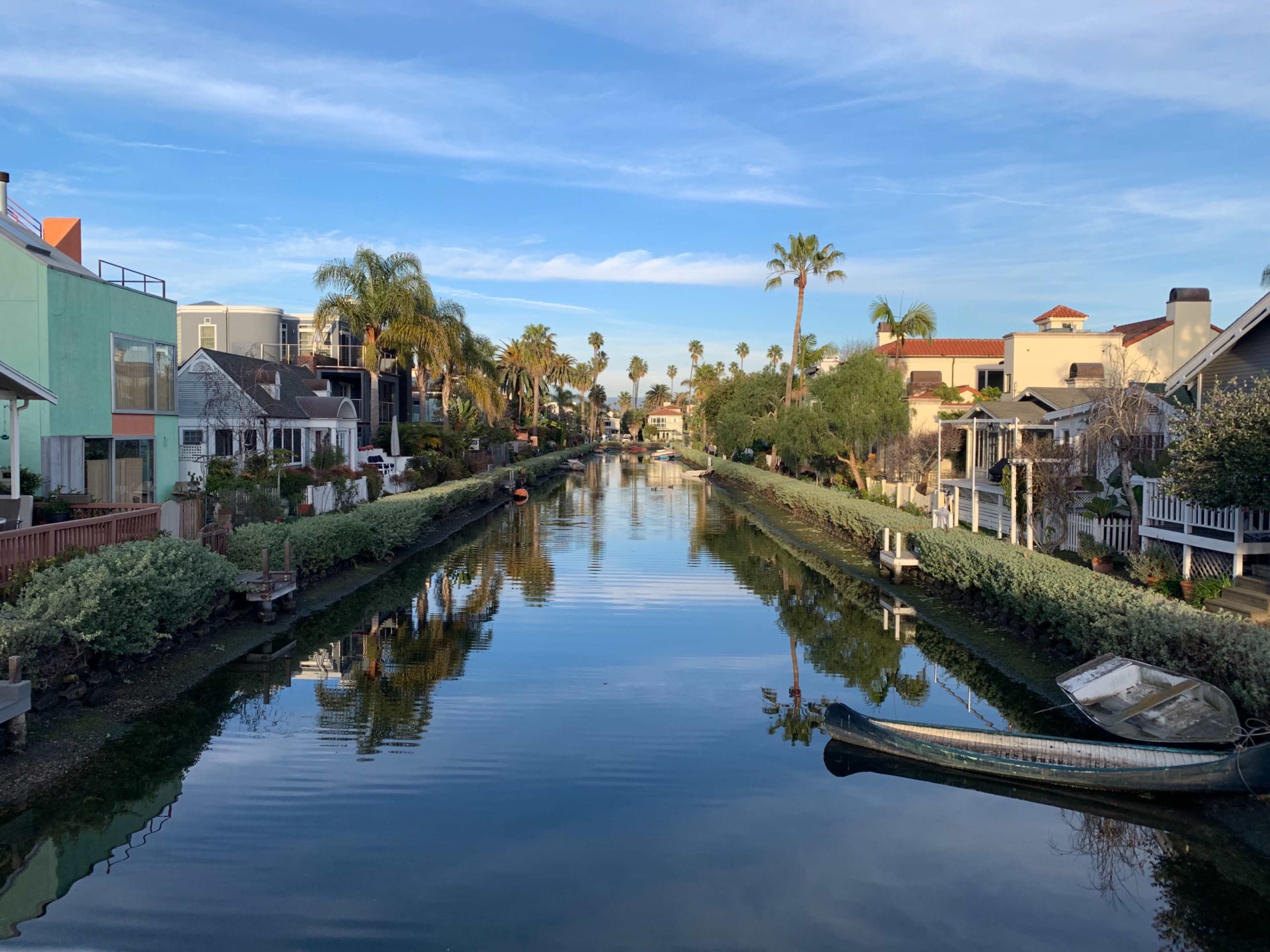 The image shows a calm canal lined with palm trees and residential houses reflecting in the water under a clear blue sky.
