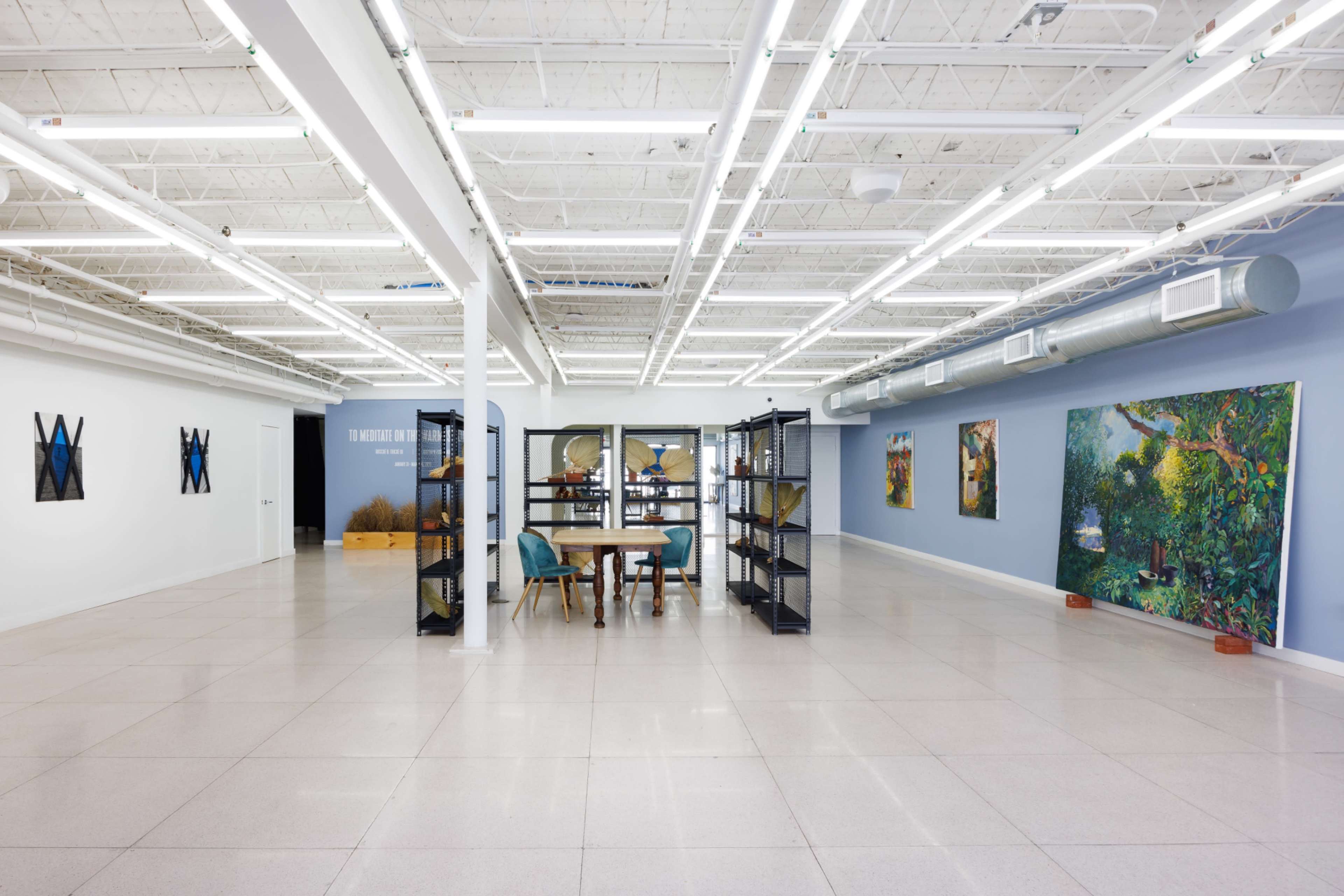 The image shows a spacious, well-lit contemporary gallery featuring a wooden table and chairs in the center, flanked by artwork displayed on the walls.