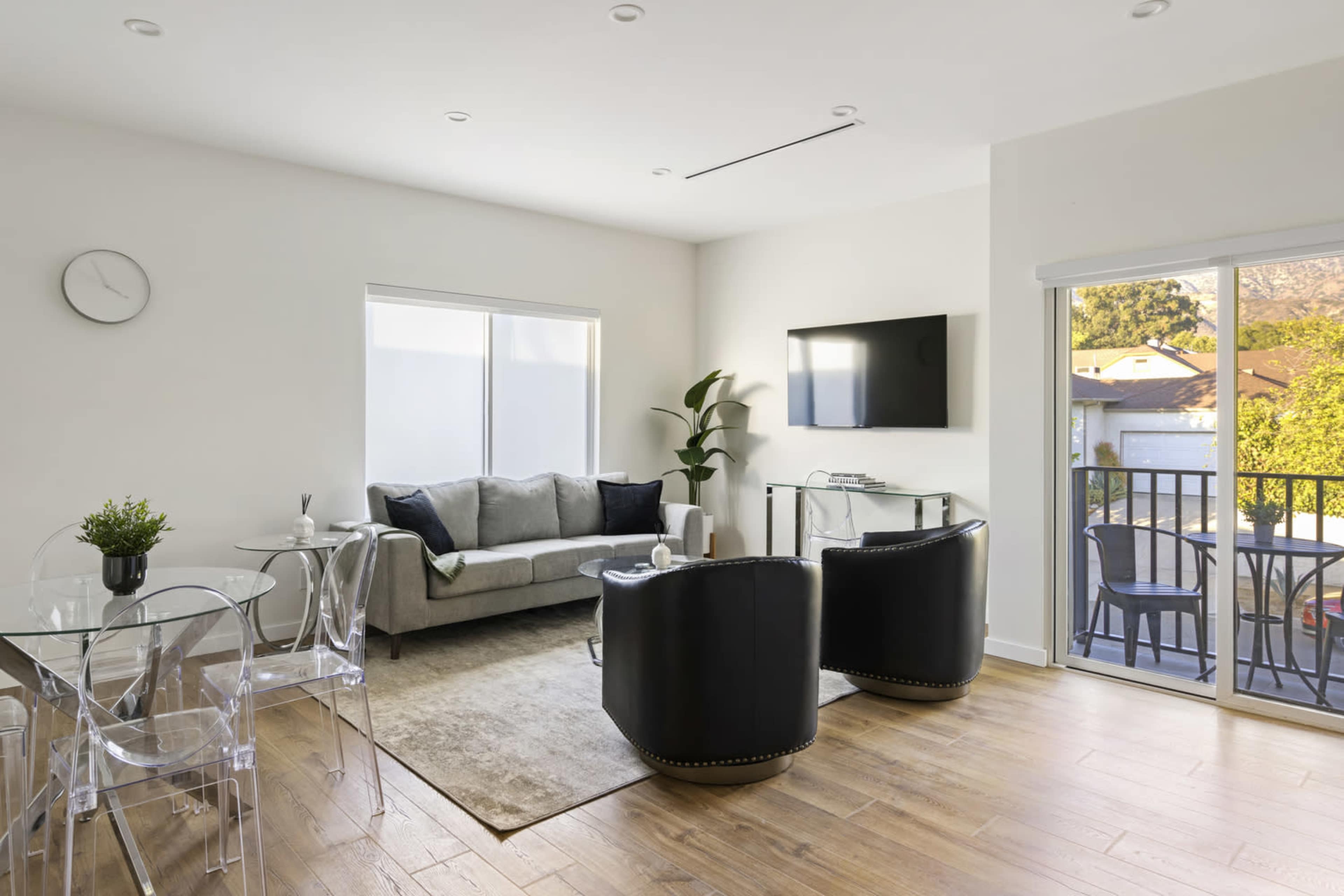 The image shows a modern living room featuring a gray sofa, two black chairs, a dining table with clear chairs, and large windows that provide natural light.