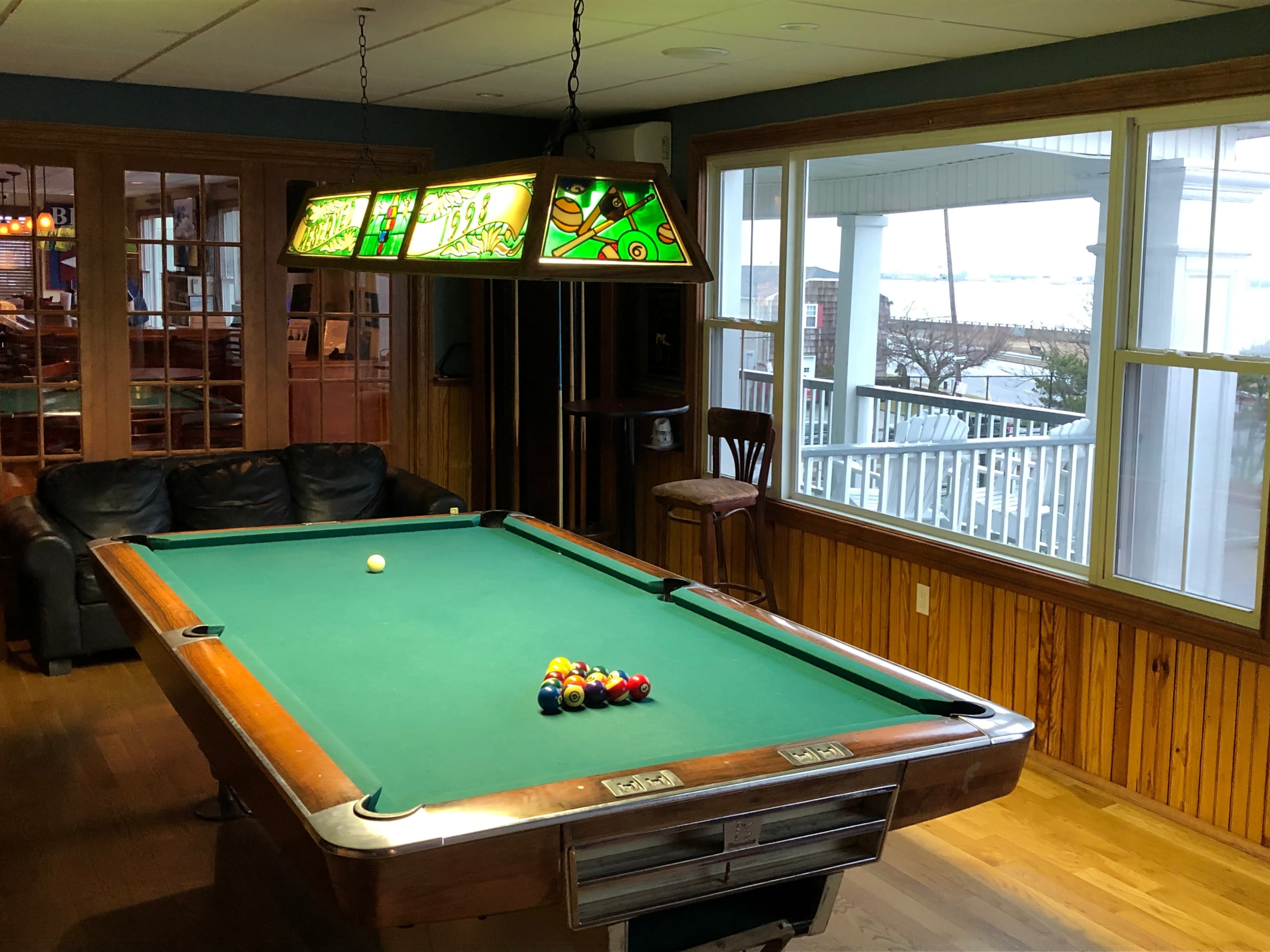 A pool table with a set of billiard balls arranged on it is located in a room with large windows overlooking a body of water.