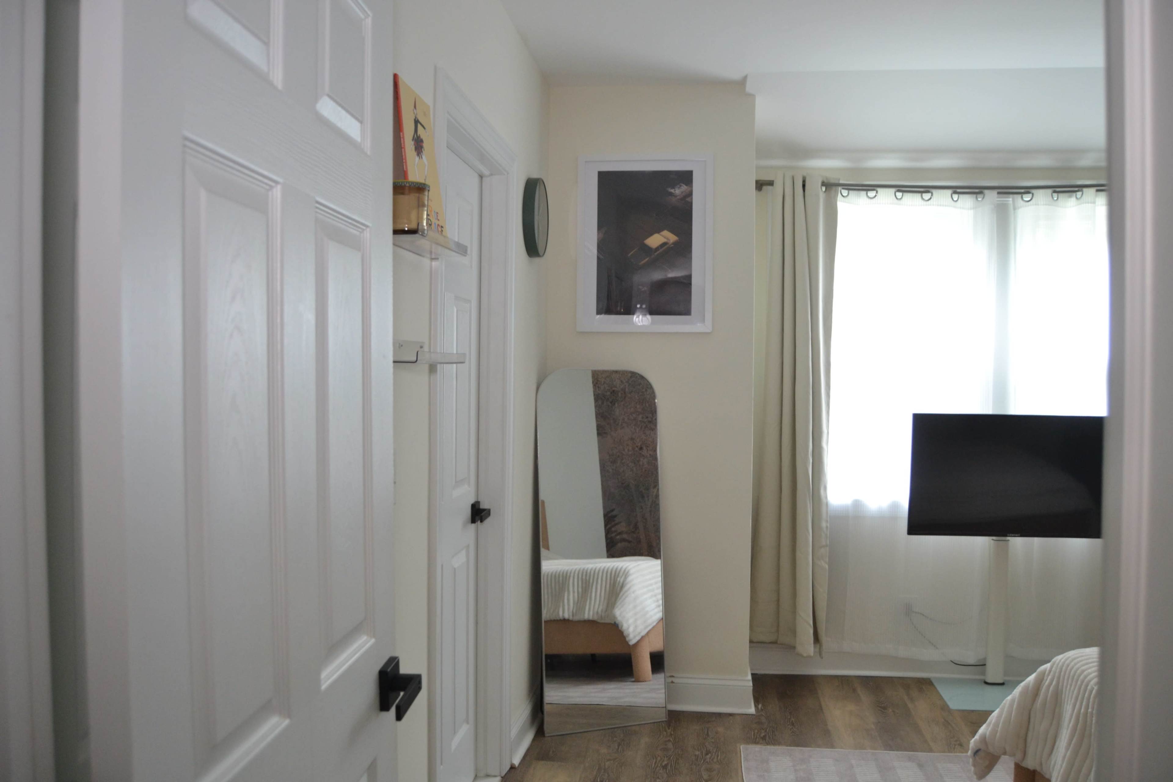 The image shows a well-lit room with a door on the left, a mirror and a television stand opposite a window with sheer curtains.