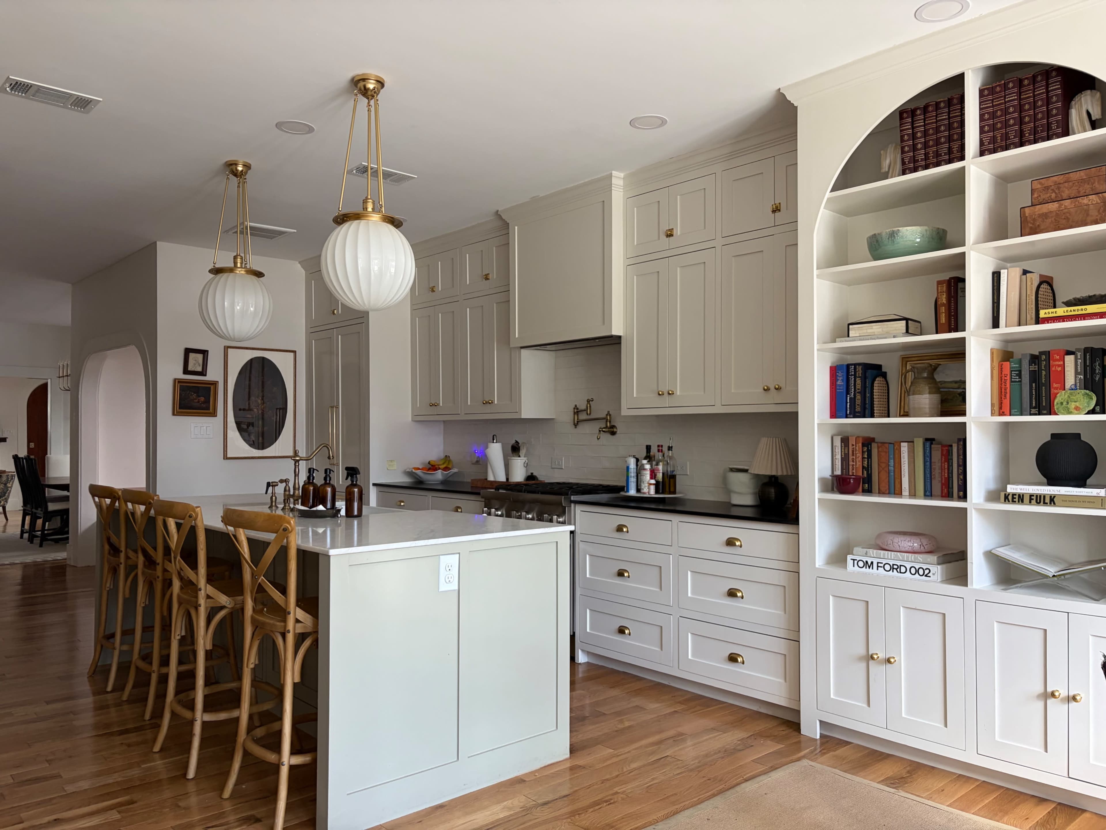 The image shows a modern kitchen with light-colored cabinetry, a central island with seating, and built-in bookshelves displaying various items and books.