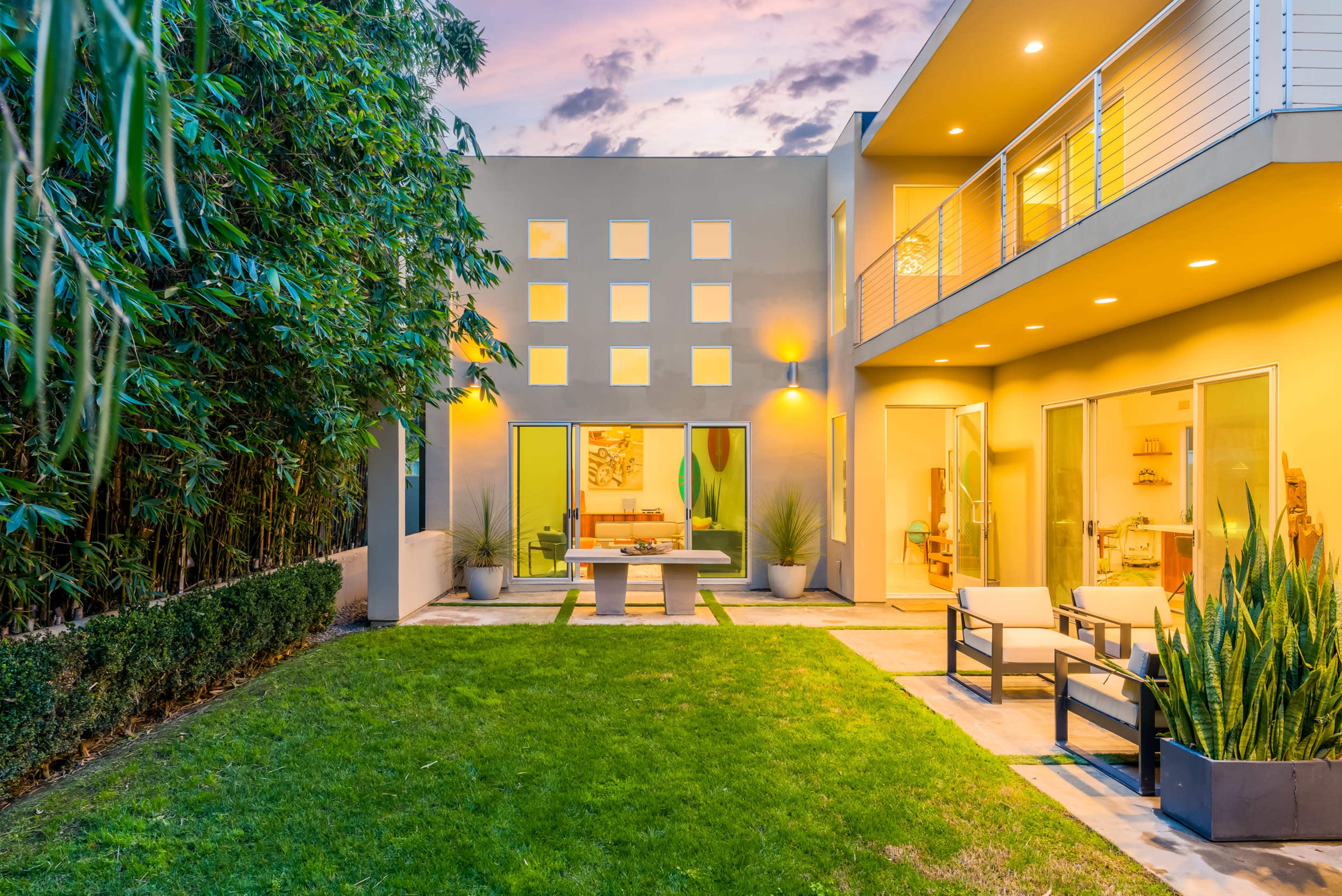 A modern two-story house features large glass doors opening to a landscaped yard with a seating area and a stone table, surrounded by greenery.