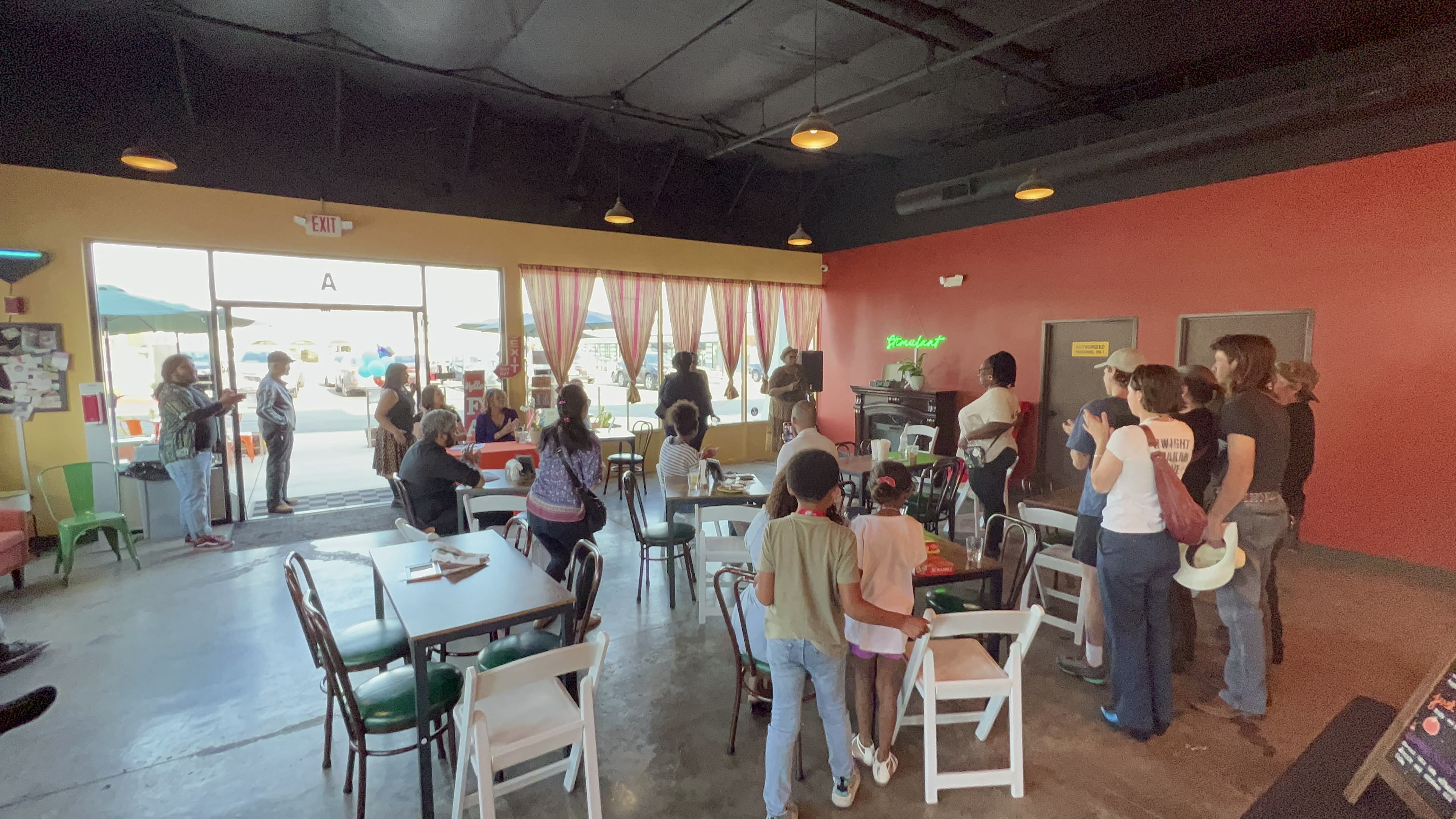 A group of people stands in line at a brightly colored restaurant, with tables and chairs arranged around the space and an open entrance leading to the outdoors.