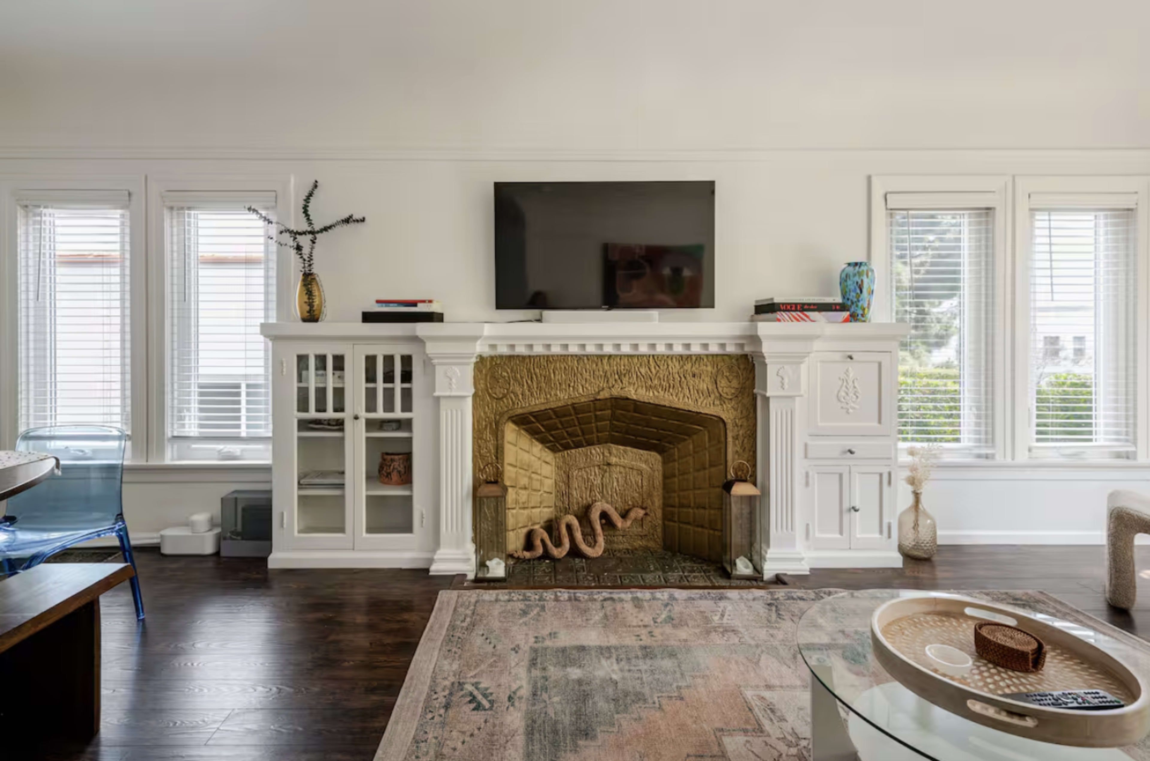 The image shows a modern living room featuring a stone fireplace with a television above it, surrounded by large windows and wooden flooring.