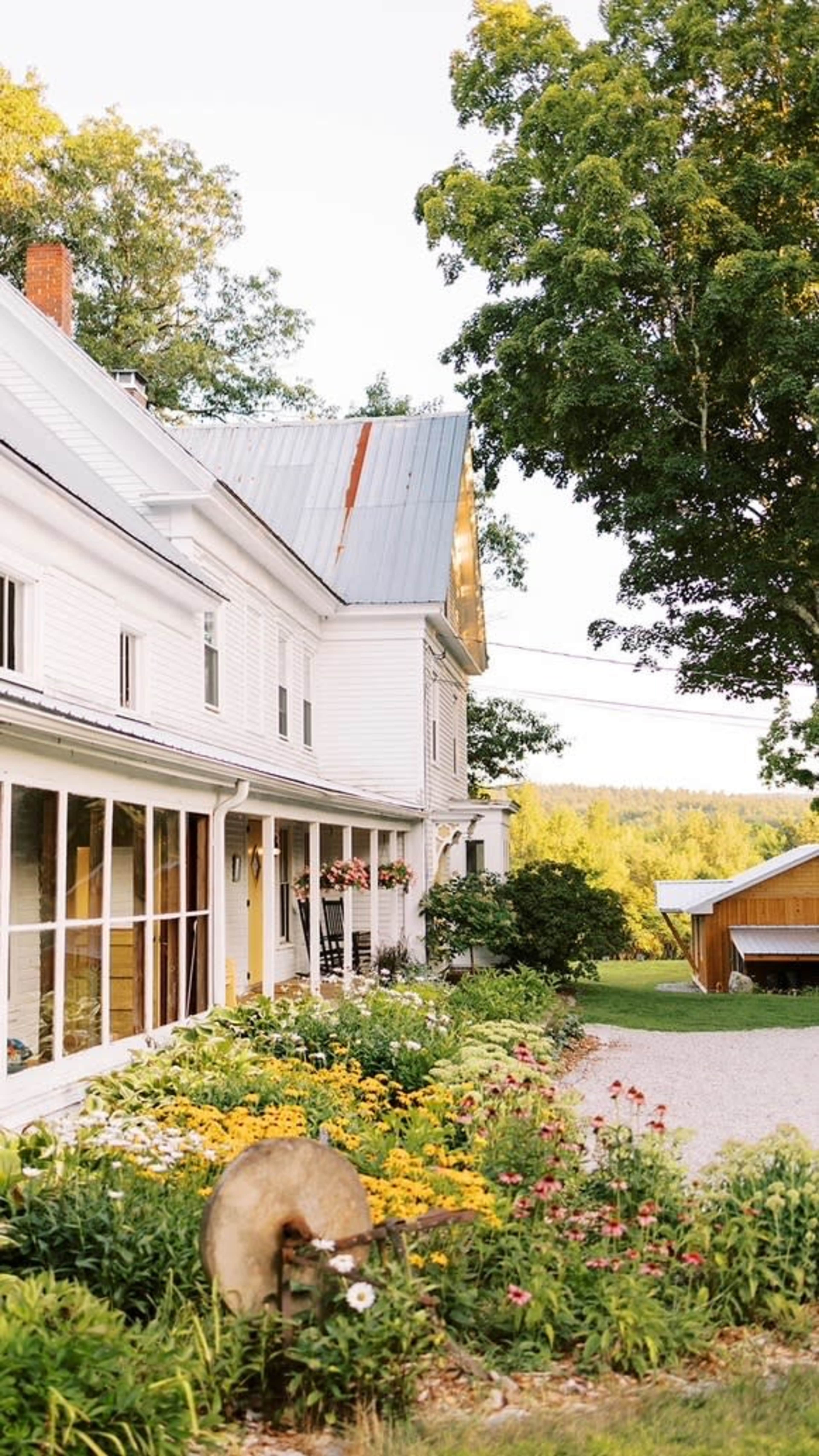 A white farmhouse with a metal roof is set behind a colorful flower garden and gravel driveway, surrounded by trees in a rural landscape.
