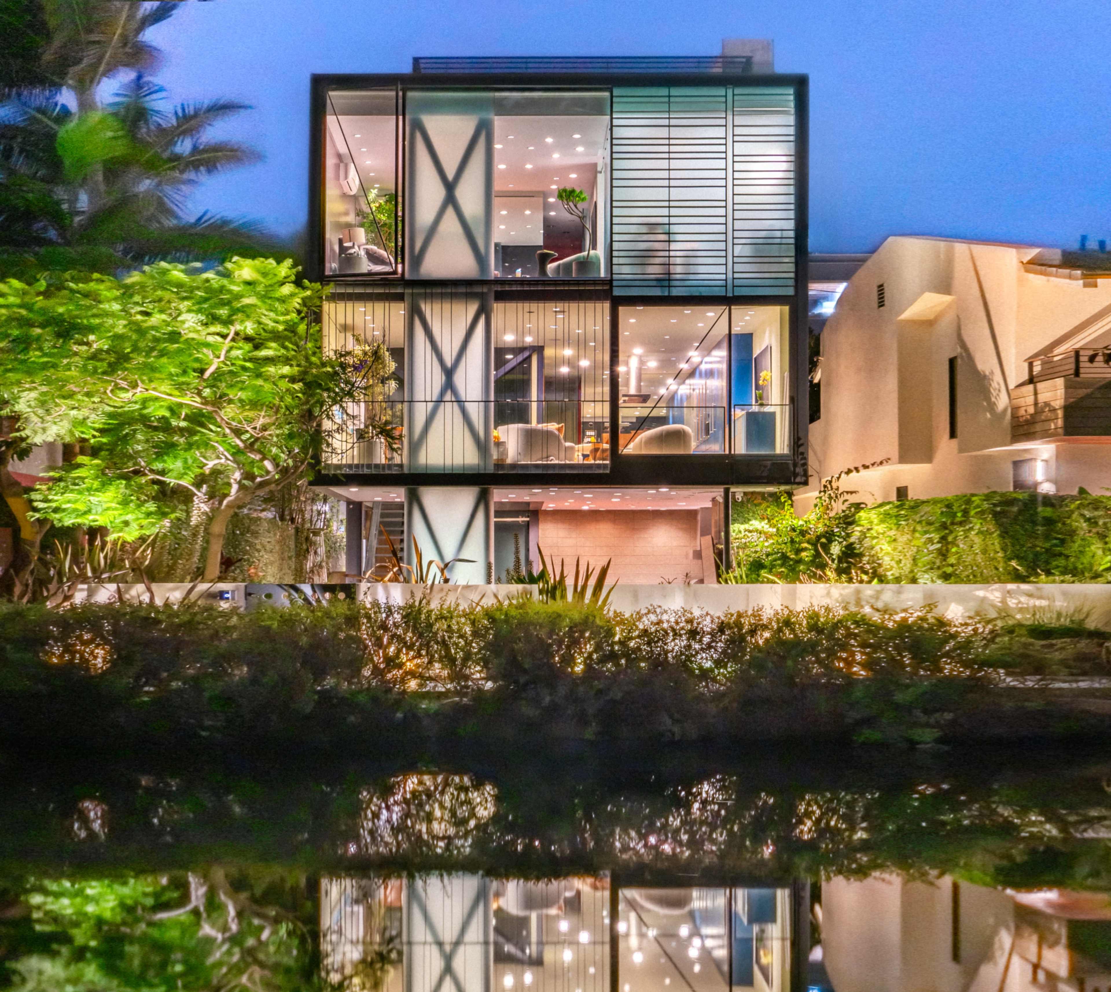 A modern three-story house with large windows and a reflective surface stands beside a body of water, surrounded by greenery.