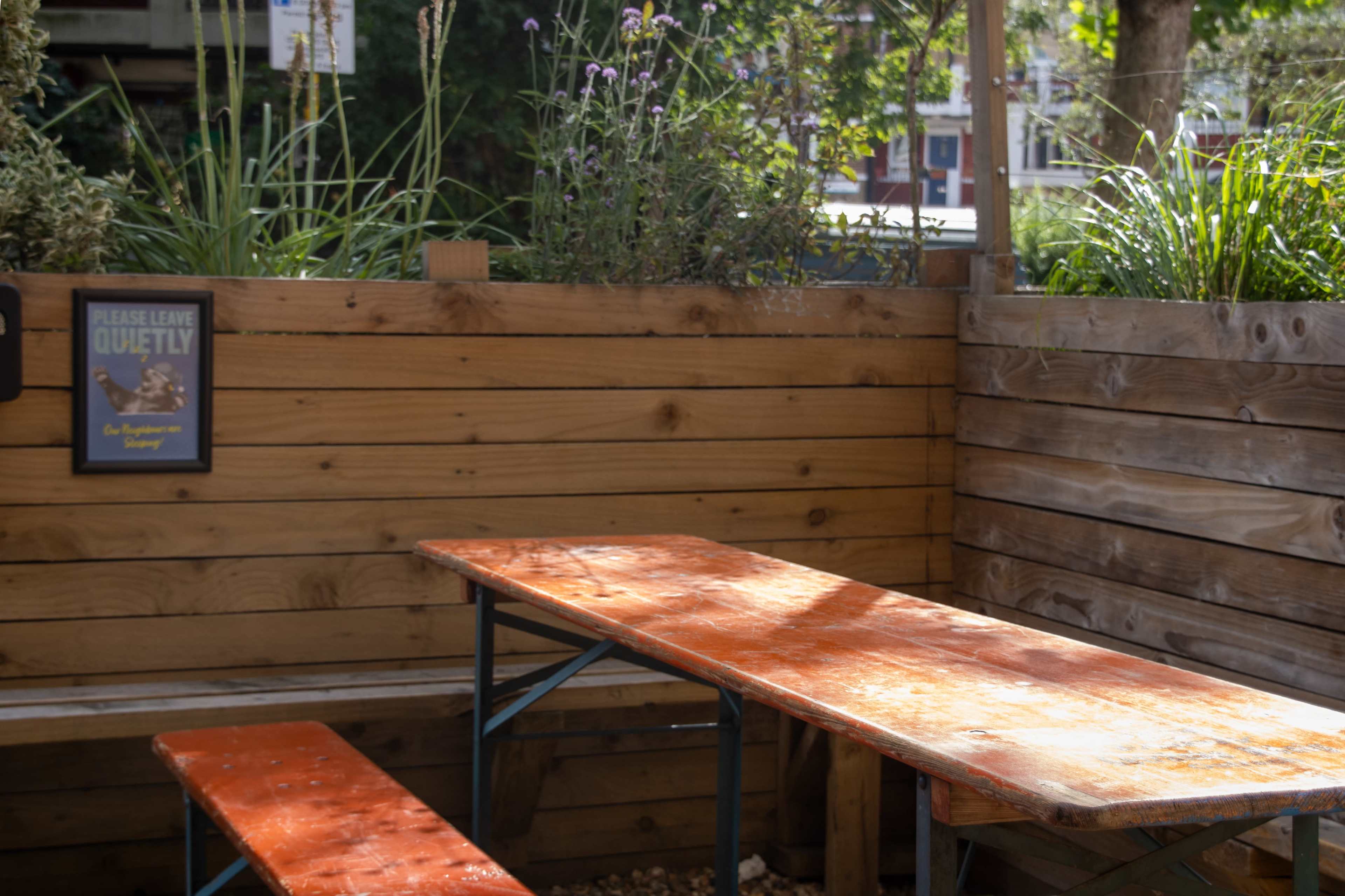 A wooden table with a bench is set in a garden area surrounded by greenery and a sign that says "Please Leave Quietly."