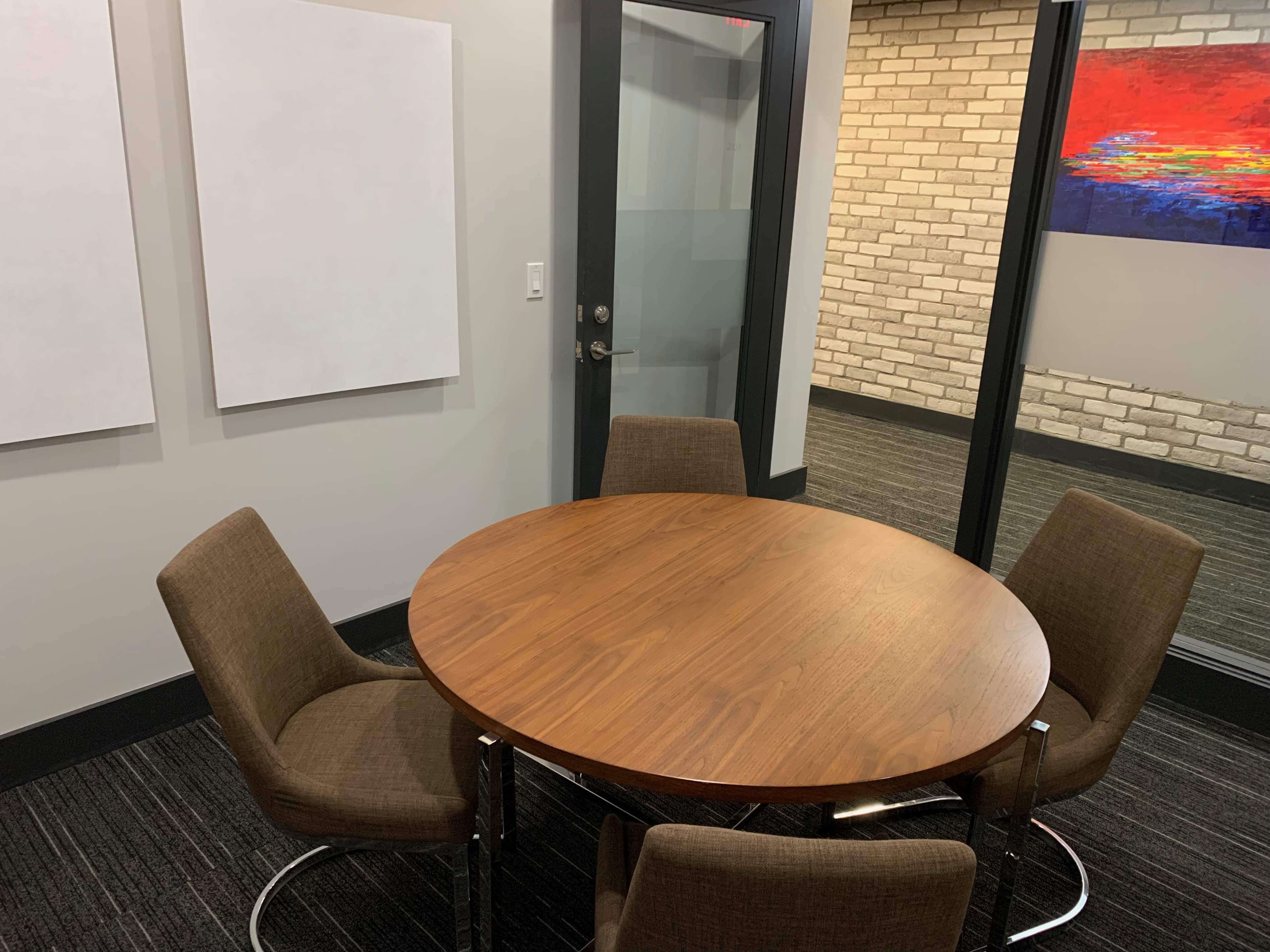 A round wooden table with four chairs is positioned in a small meeting room featuring two blank white wall spaces and a glass partition.