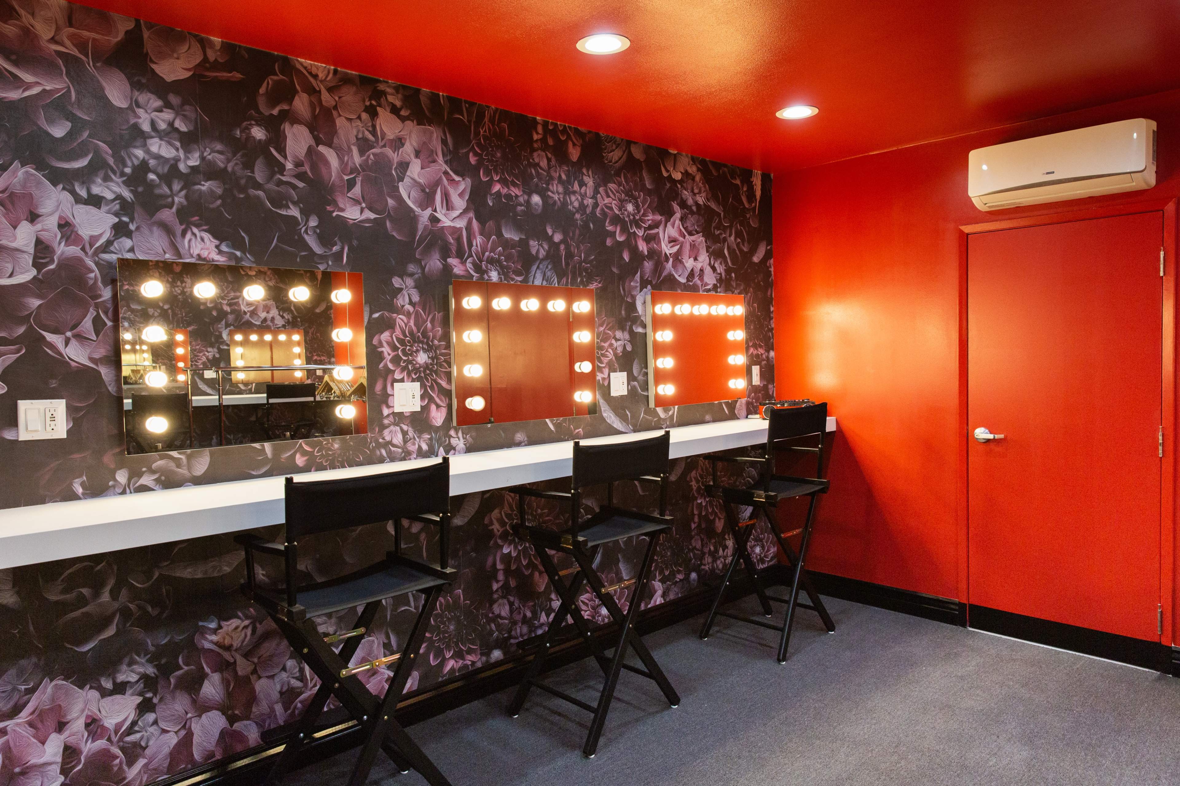 A brightly colored dressing room featuring three illuminated mirrors and black folding chairs against a floral-patterned wall.