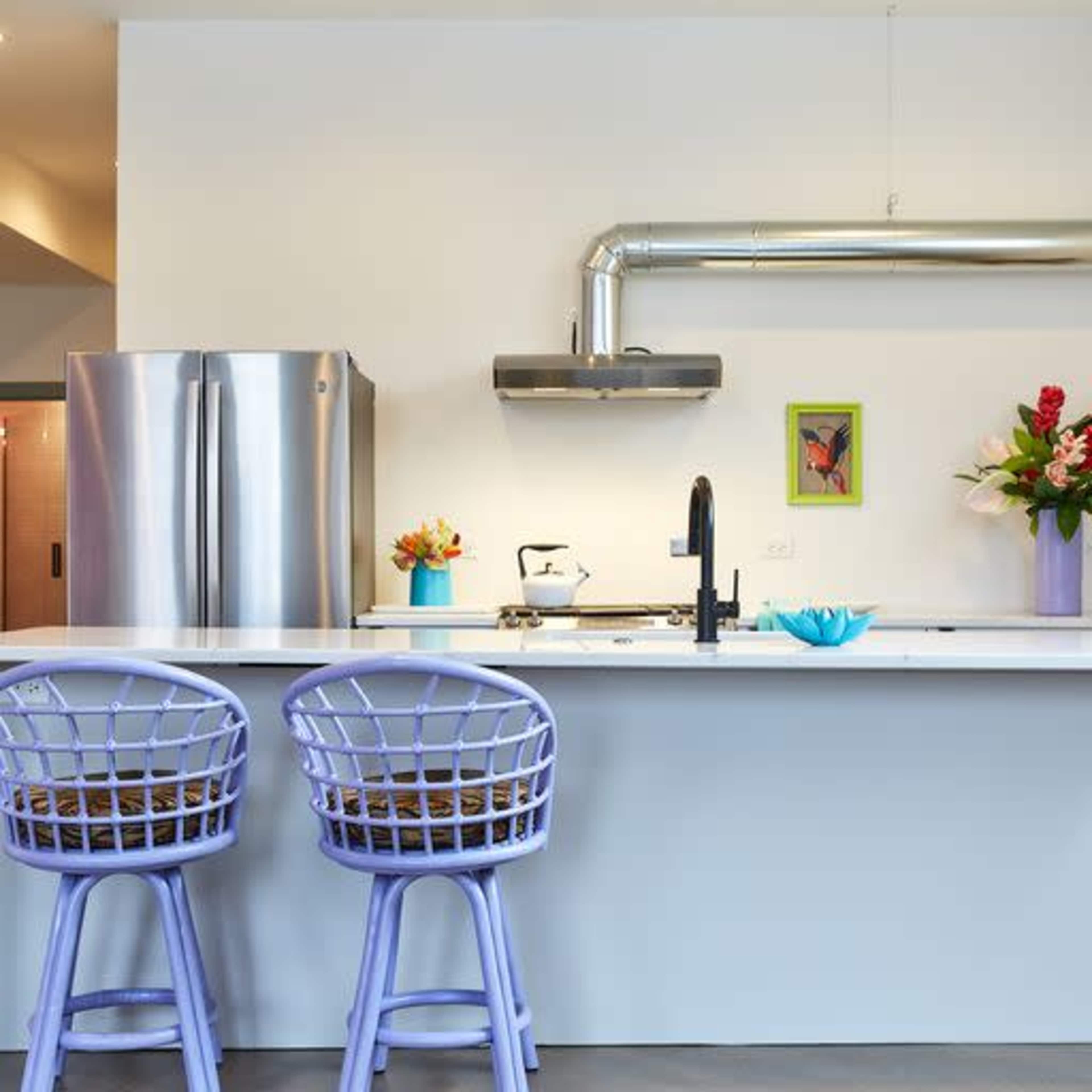 A modern kitchen features a stainless steel refrigerator, a white countertop with decorative items, and three purple bar stools.