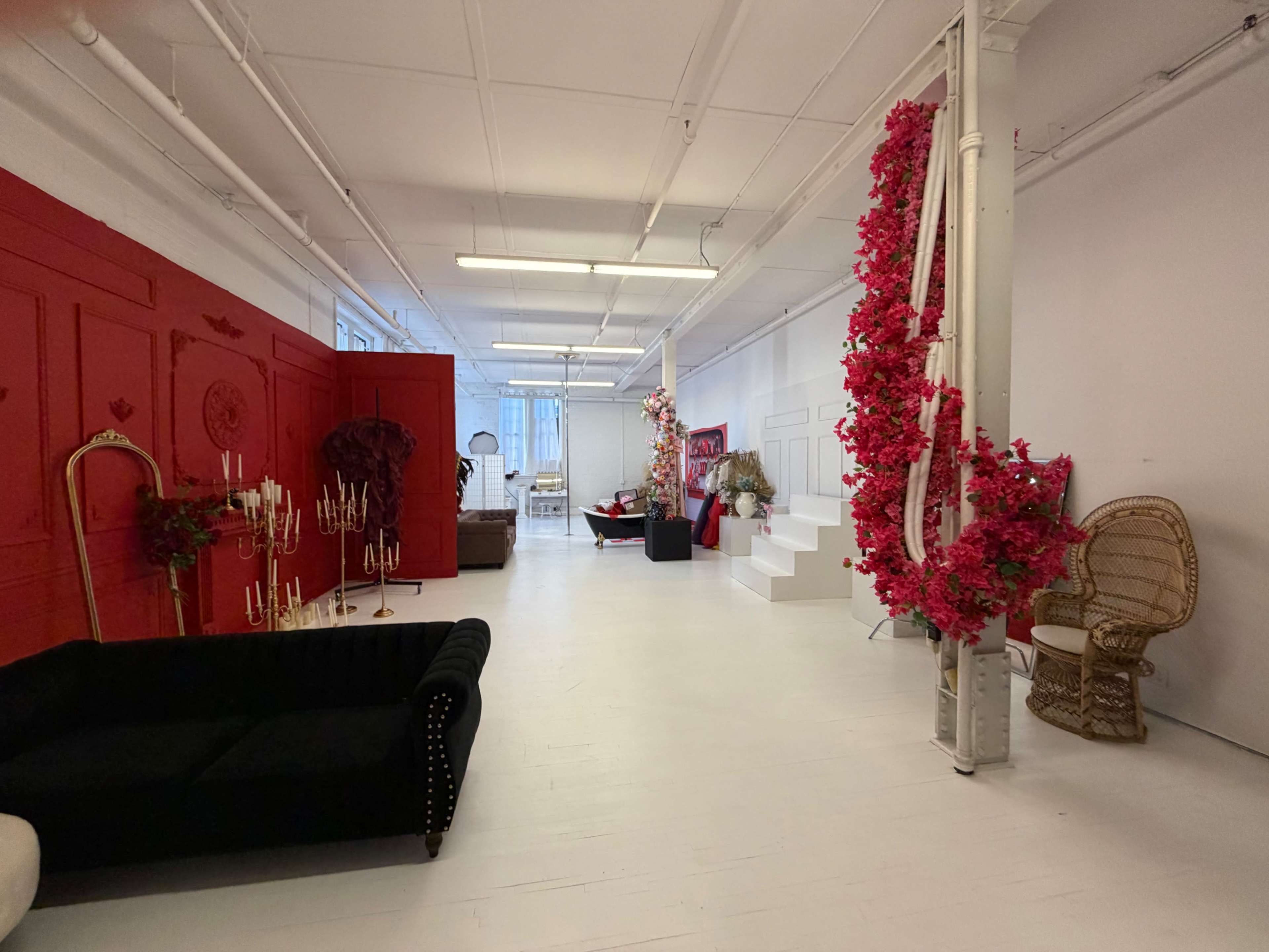 The image depicts a spacious interior with white floors and walls, featuring red accent walls and decor, along with various furnishings including a black sofa and a decorative staircase.