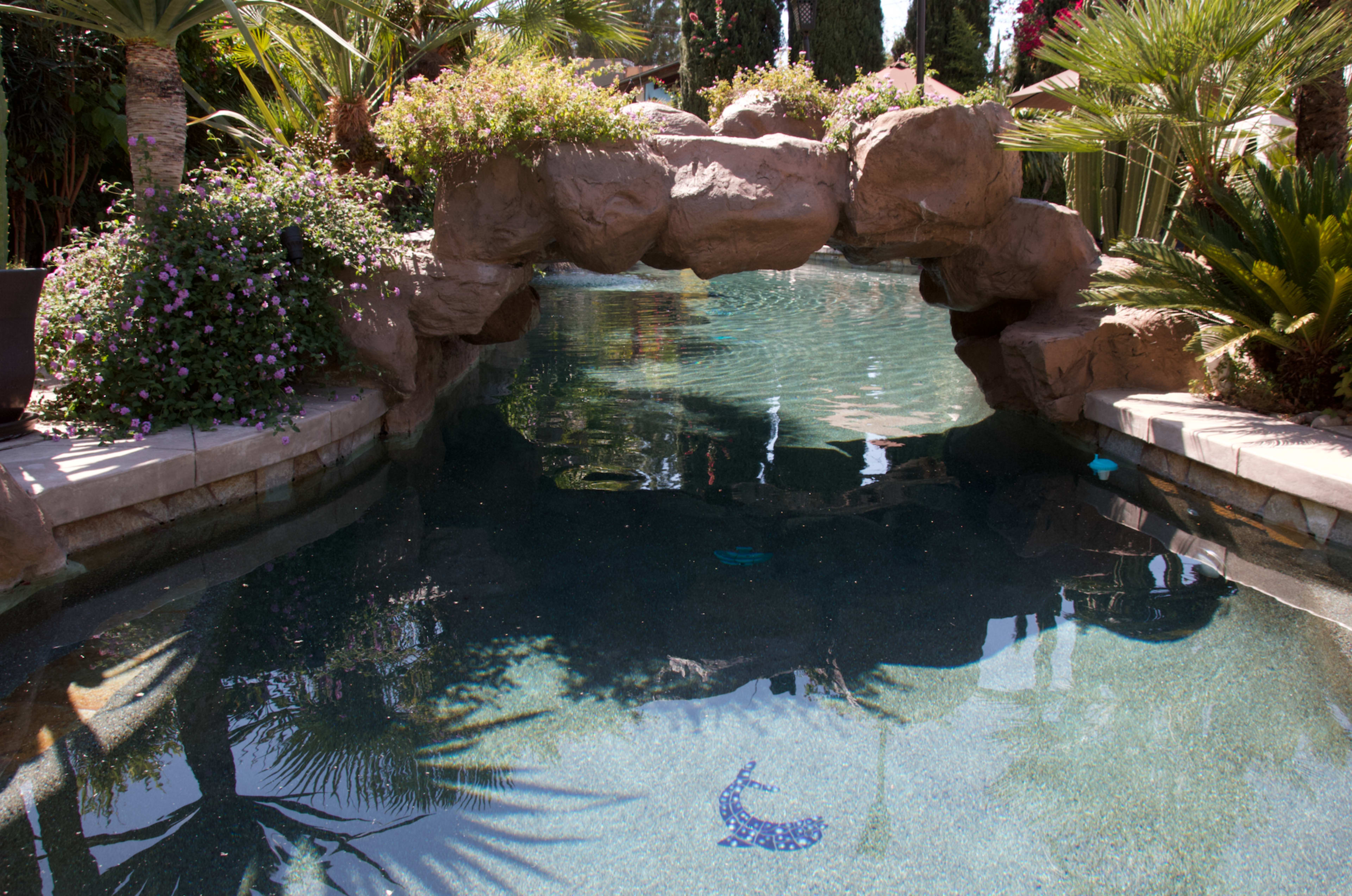 A swimming pool with a stone arch bridge surrounded by lush greenery and colorful flowers.